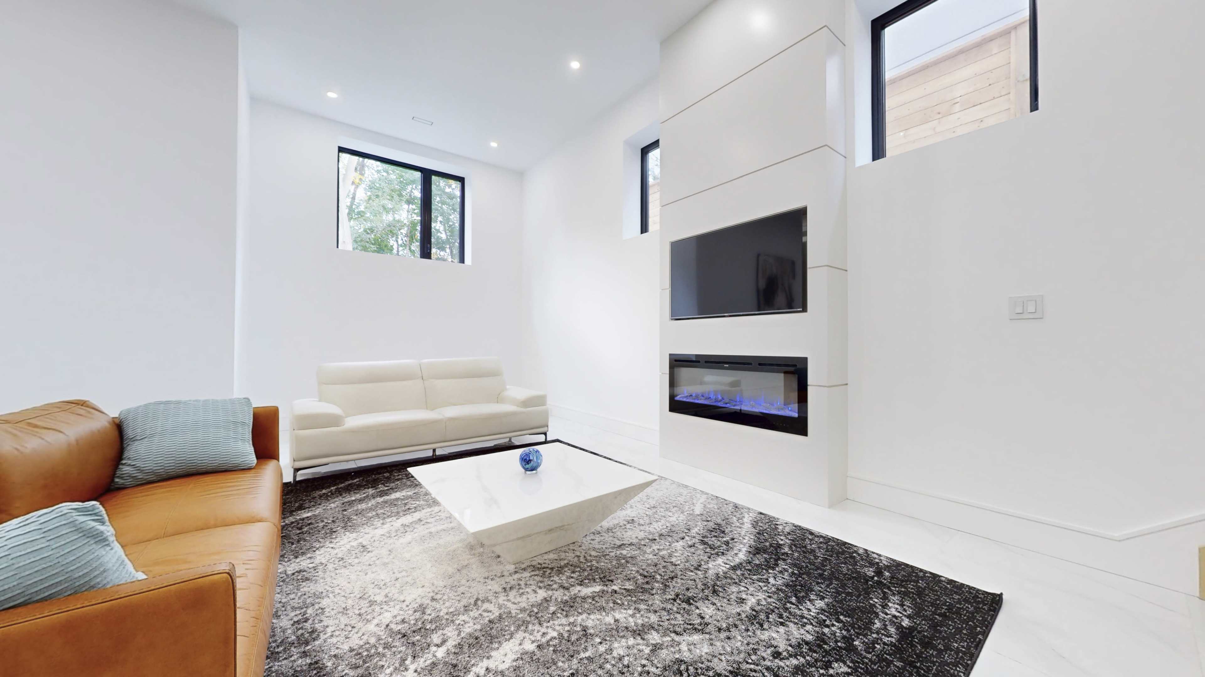 A modern living room with a white wall, a beige sofa, a flat-screen TV mounted above a sleek fireplace, and a patterned black and white rug under a coffee table.