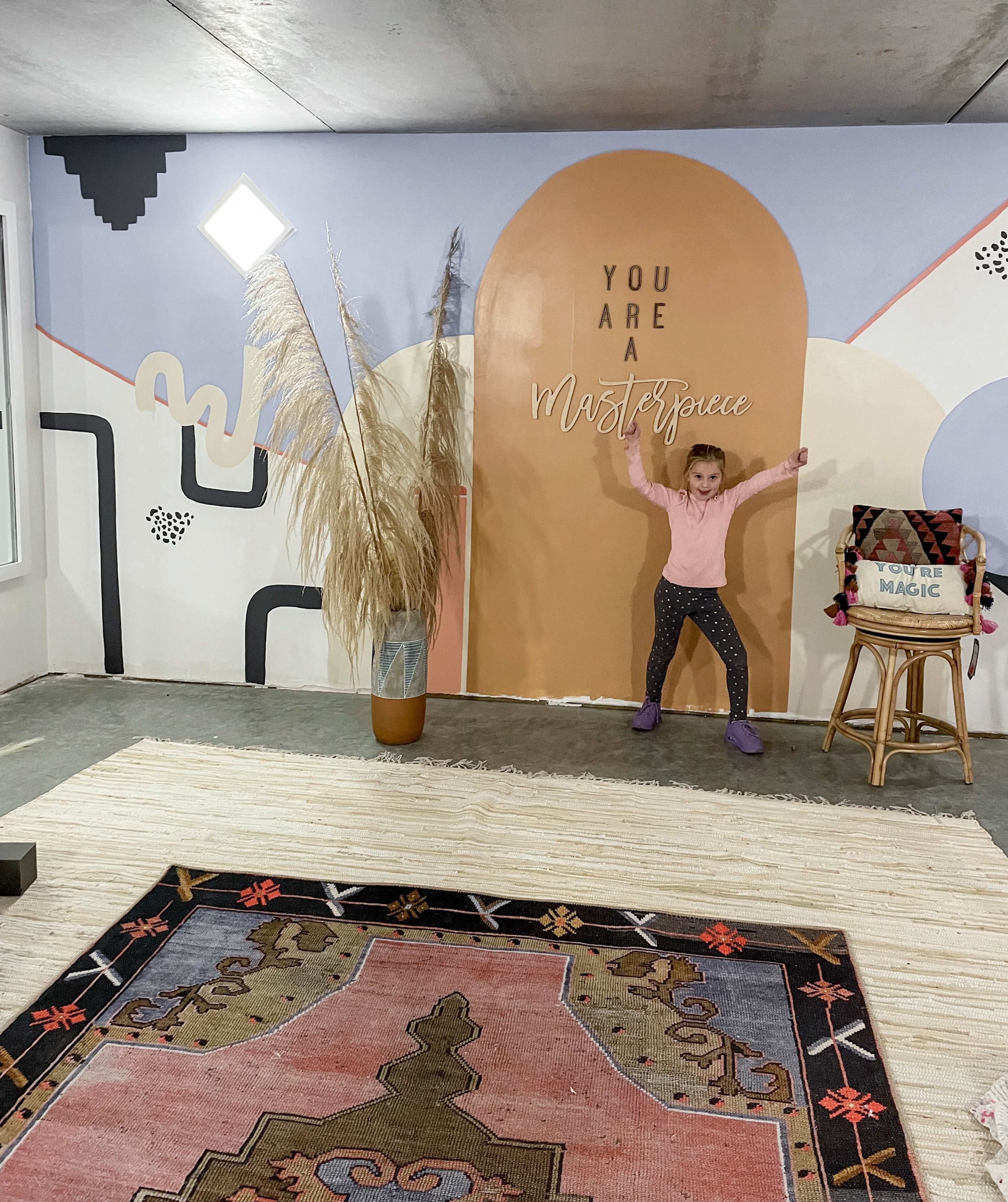 A child stands in front of a large mural that reads "You Are A Masterpiece," with decorative plants and a colorful rug on the floor.