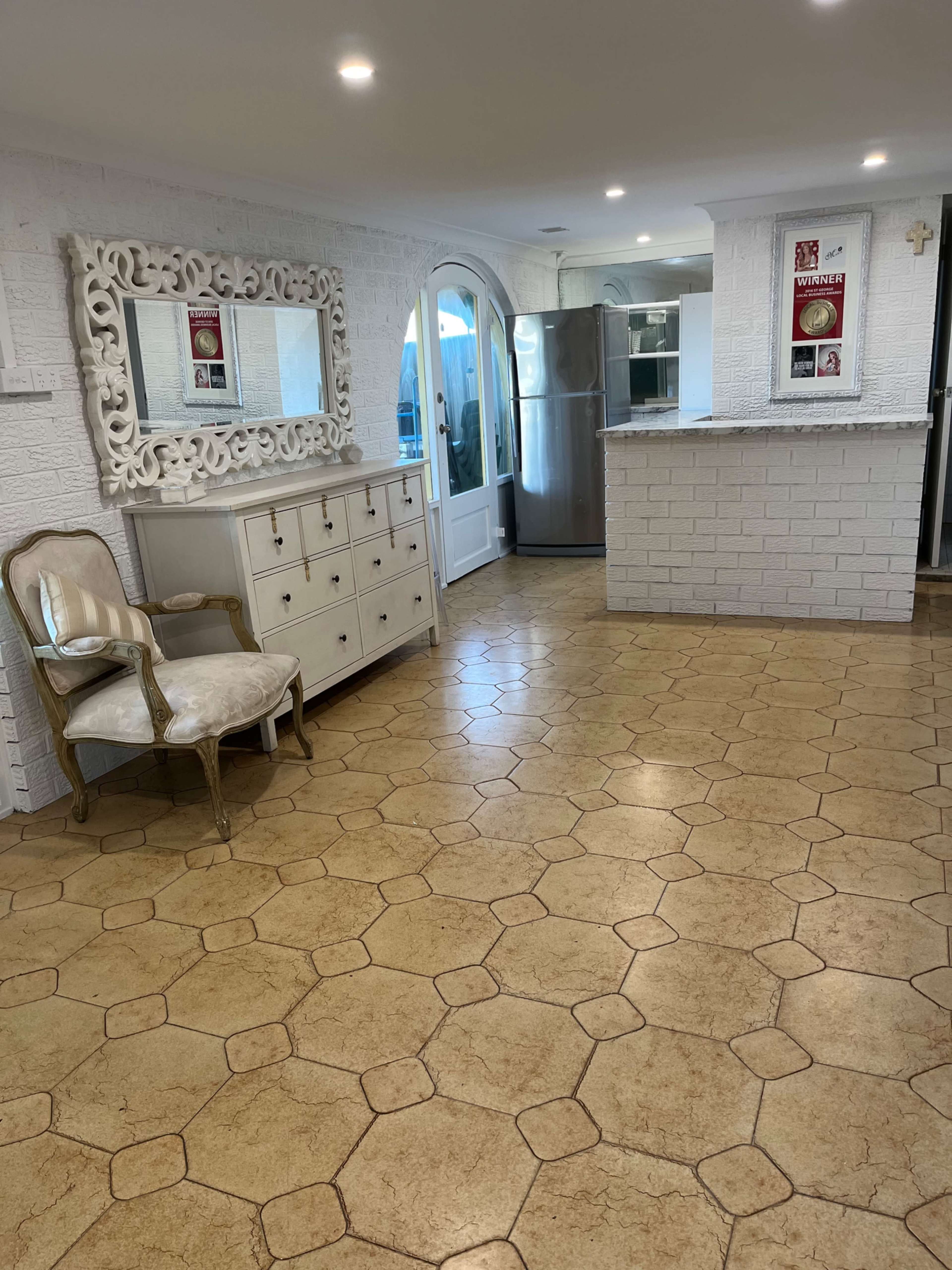 The image shows an interior space featuring a light-colored tiled floor, a decorative mirror on the wall, a white cabinet with drawers, and a small chair beside a kitchen area with a stainless steel refrigerator.