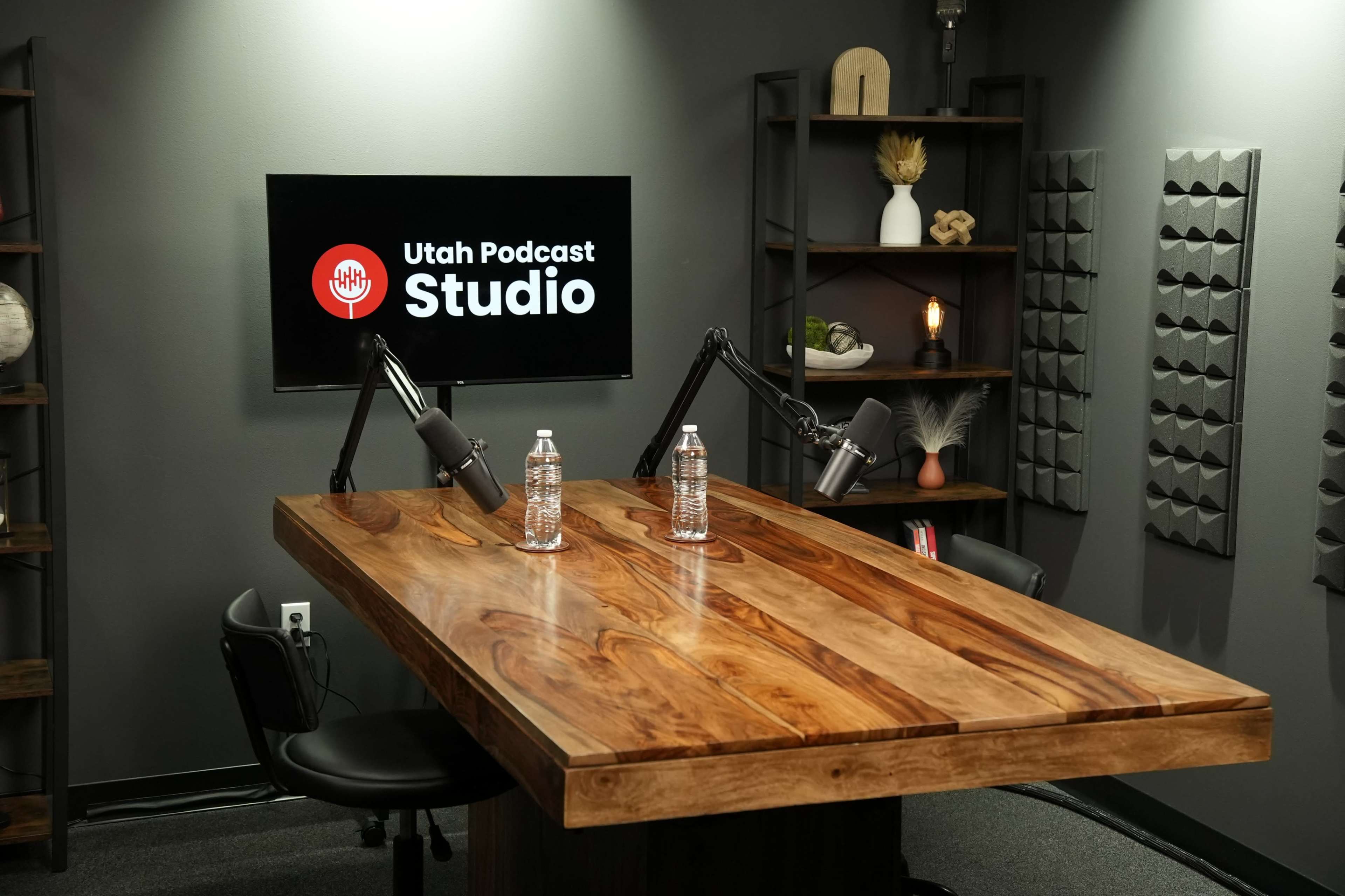 The image shows a podcast recording studio featuring a large wooden table, two water bottles, and a monitor displaying the words "Utah Podcast Studio."