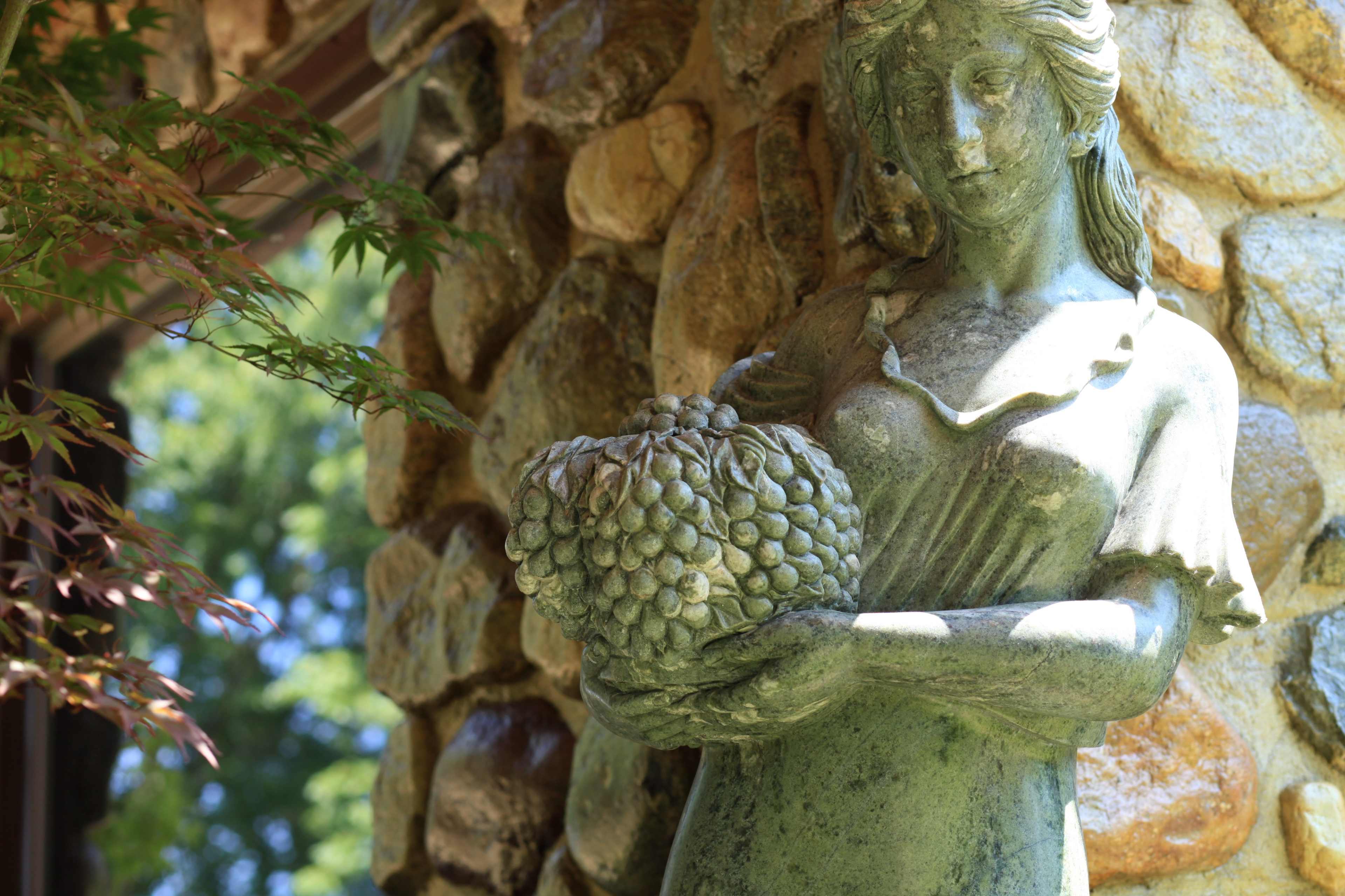 A sculpture of a woman holds a large bunch of grapes, set against a stone wall.