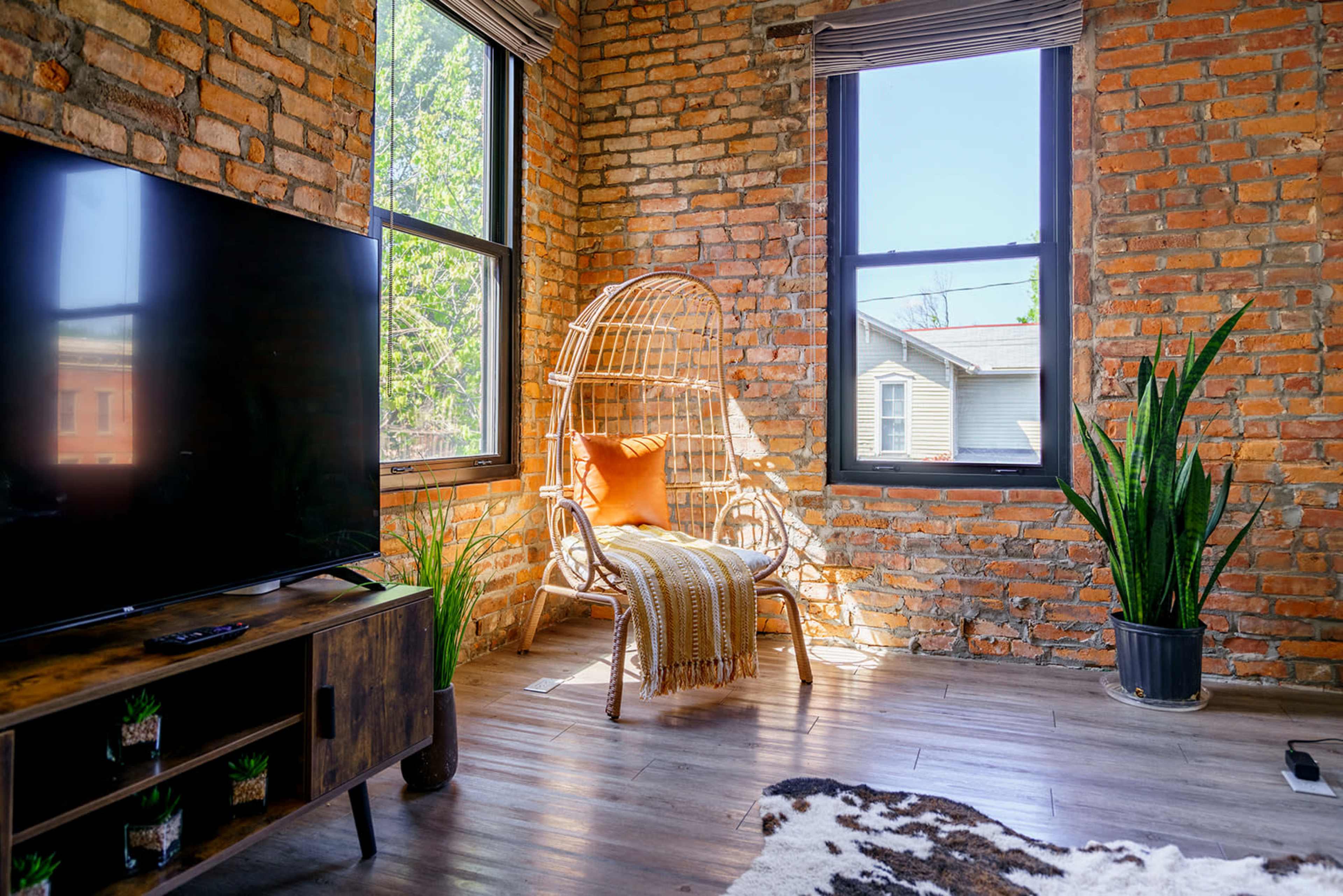 The image shows a cozy interior featuring a rattan chair, a television, and exposed brick walls with sunlight streaming through large windows.