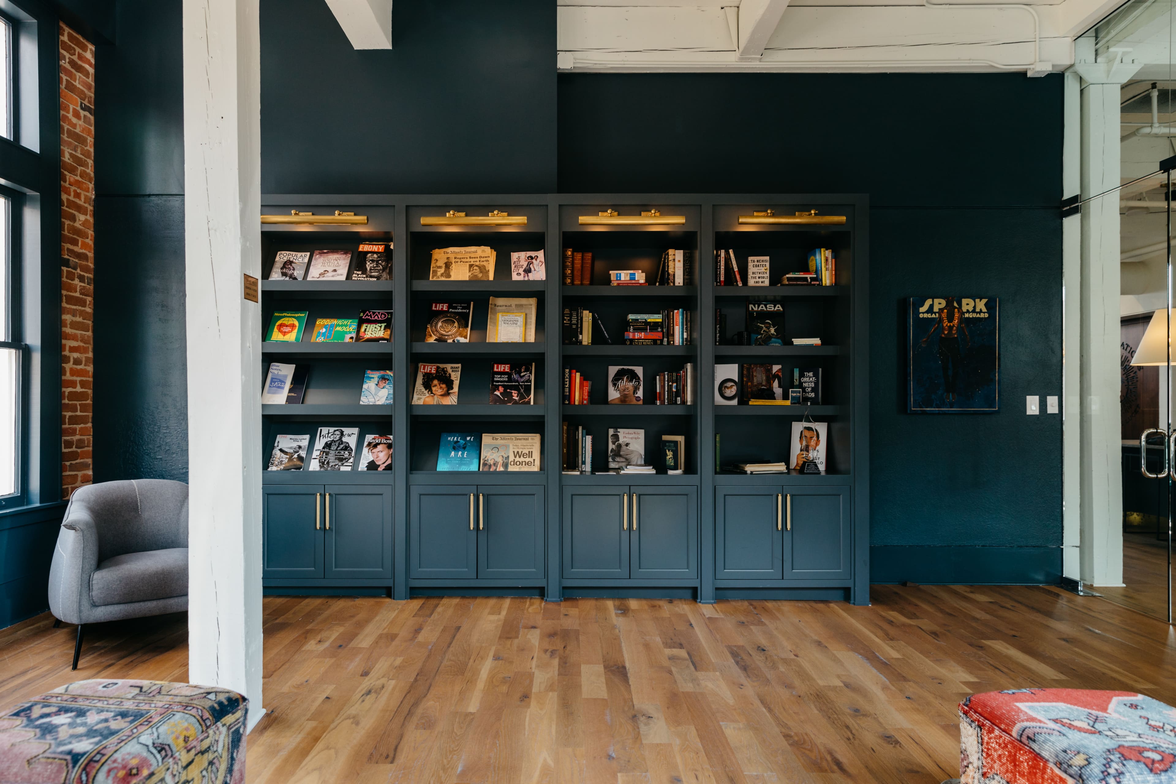 A gray bookshelf filled with books and magazines is positioned against a dark wall in a room with wooden flooring and a gray armchair nearby.