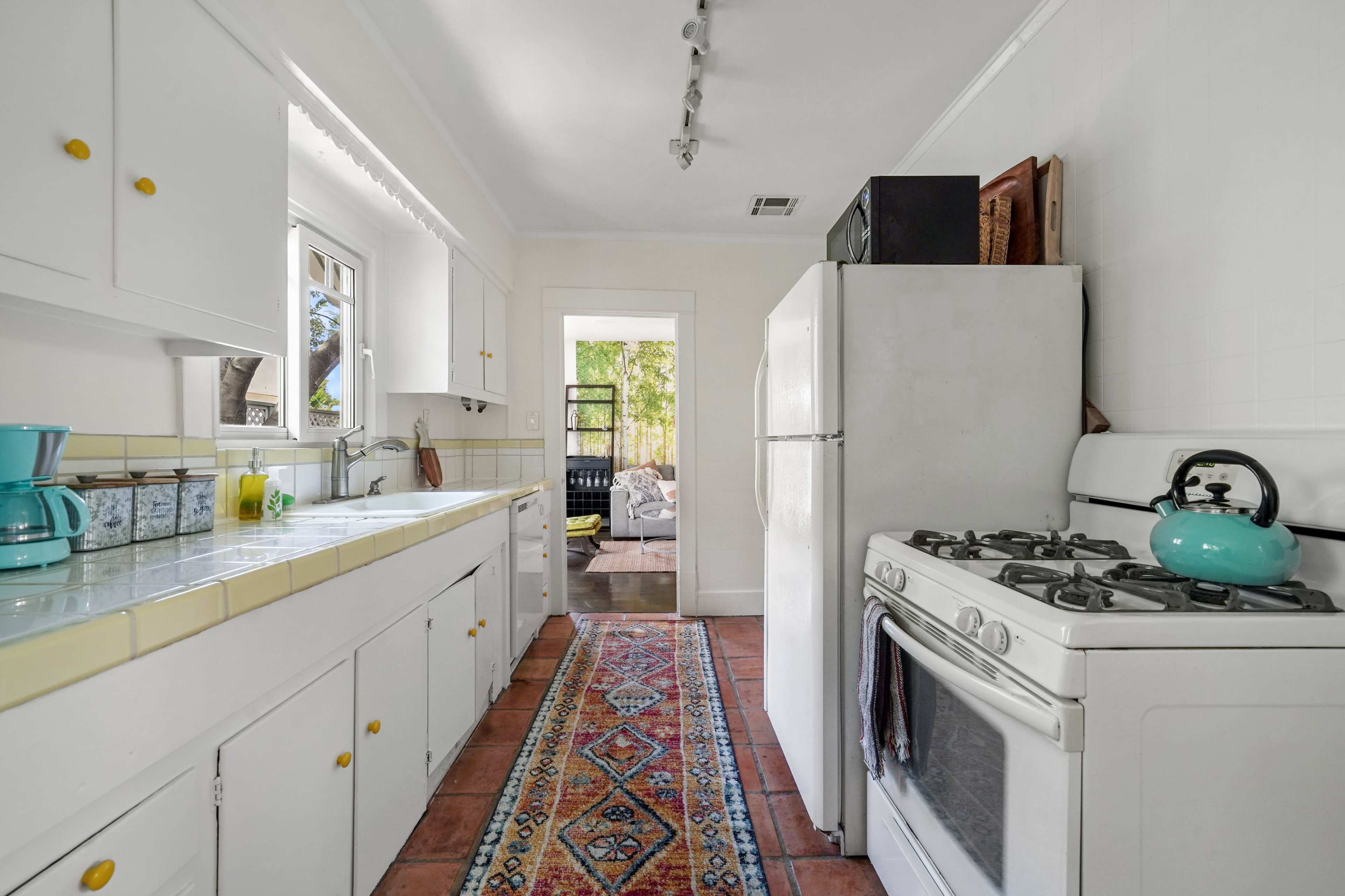 A bright kitchen features white cabinets, a white refrigerator and stove, and a colorful rug along the tiled floor.