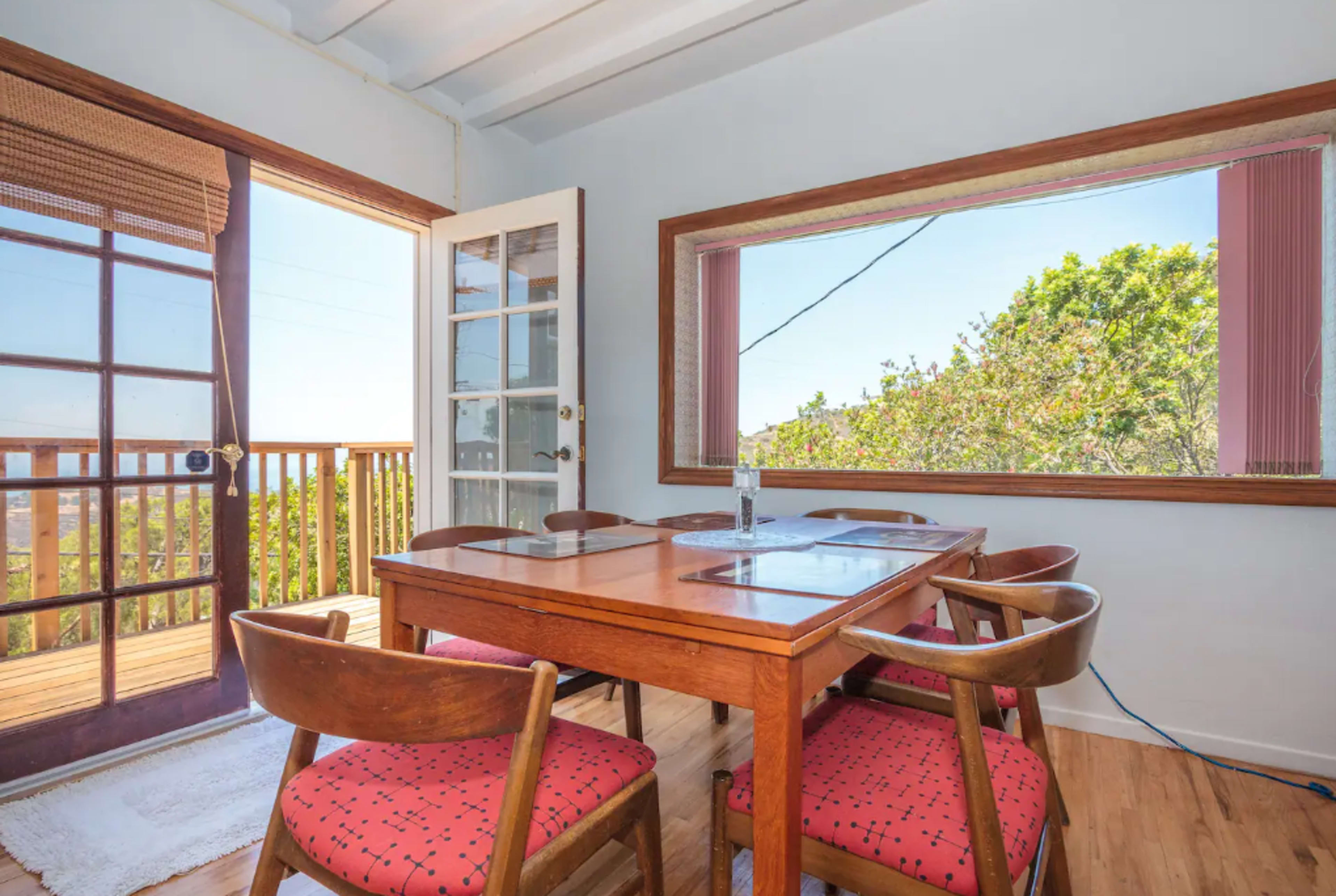 A dining area with a wooden table surrounded by four chairs, featuring a sliding glass door that opens to a patio area and a large window with a view of greenery outside.