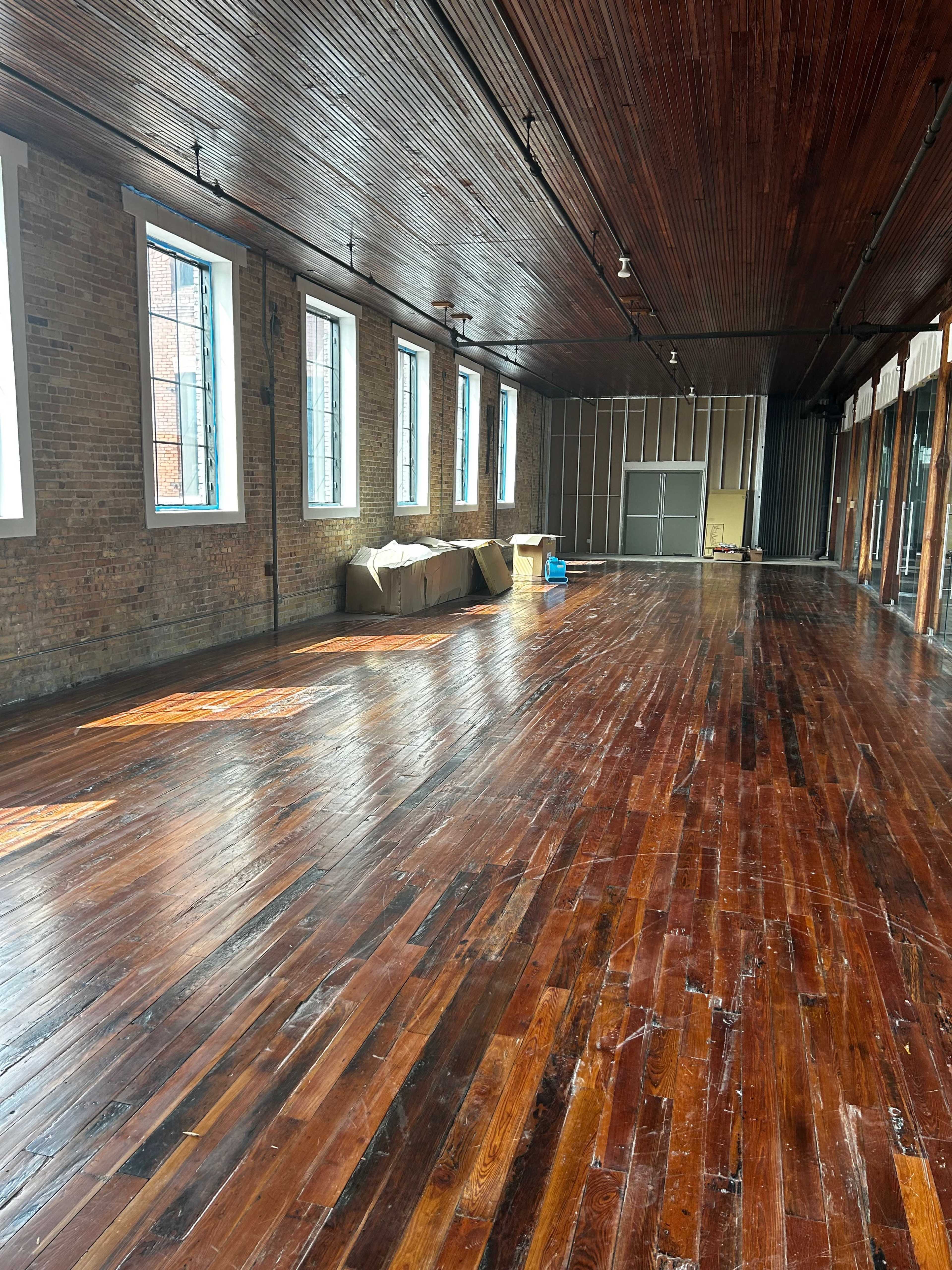 The image shows a spacious room with wooden floors, large windows, and exposed brick walls.