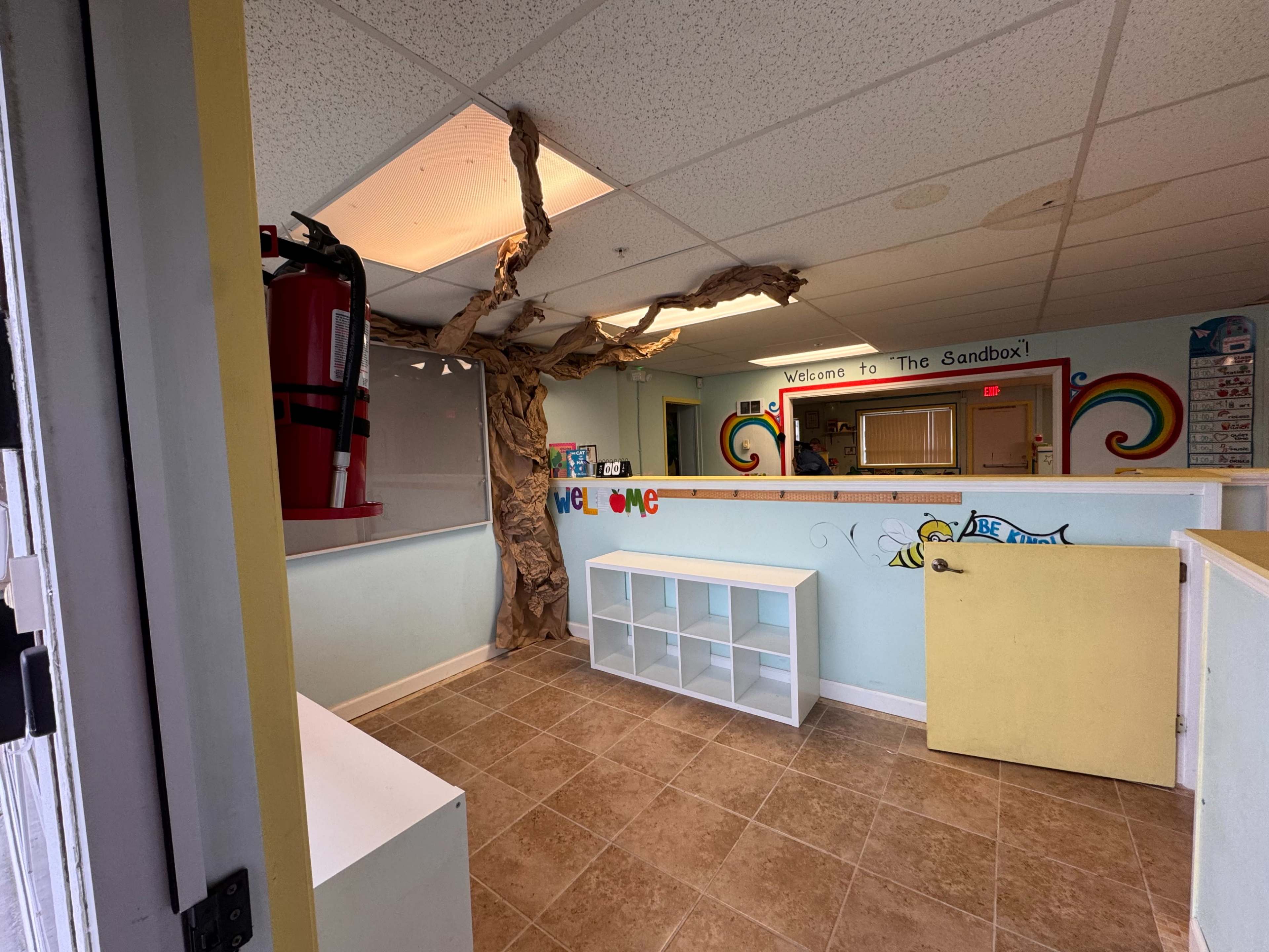 The image shows a brightly colored play area called "The Sandbox," featuring a large tree branch décor and storage shelves against pastel walls.