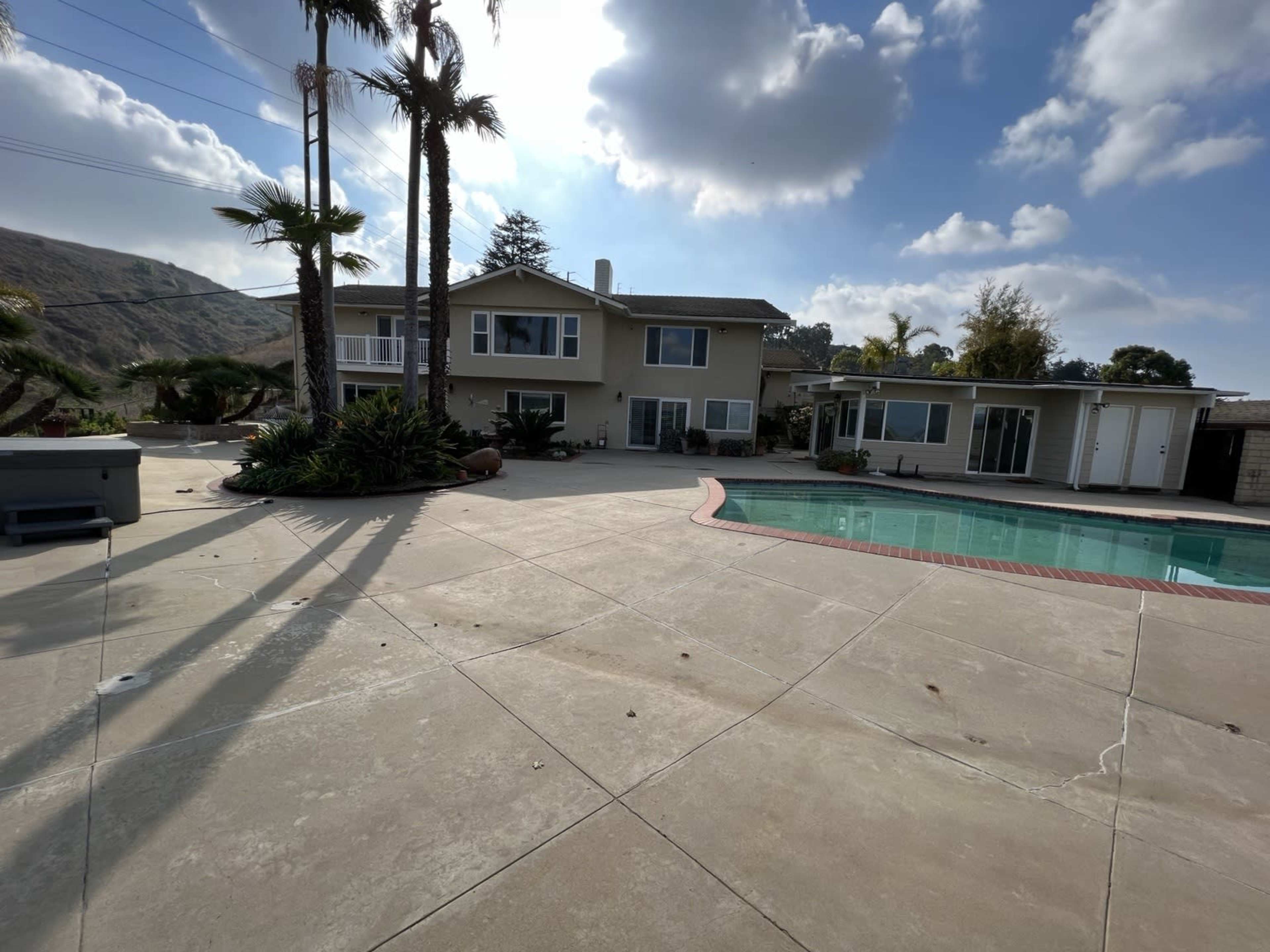 The image shows a residential property with a swimming pool, palm trees, and a large patio area under a partly cloudy sky.