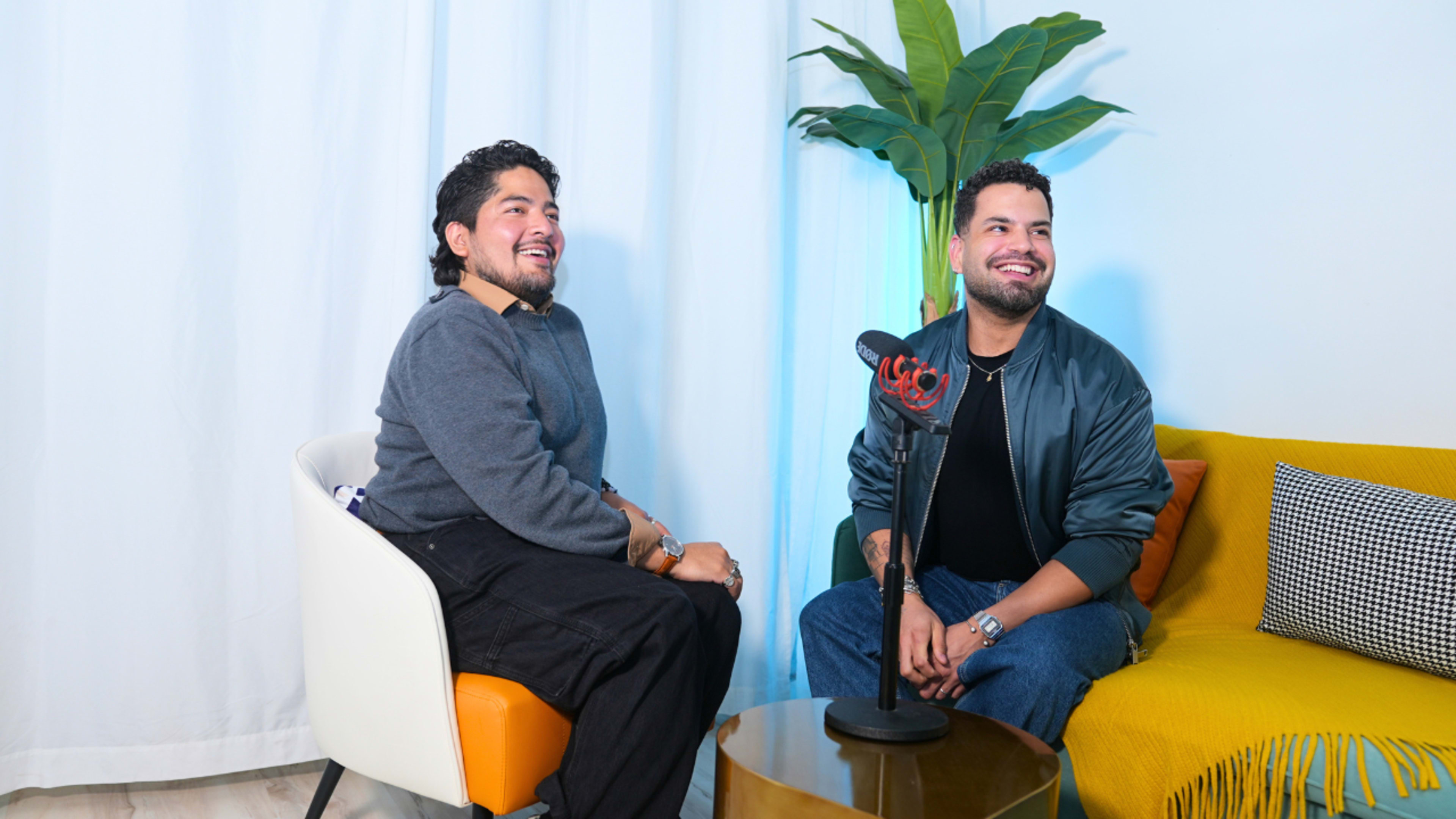 Two men sit on a colorful sofa in a well-lit room, smiling while facing a microphone on a table.
