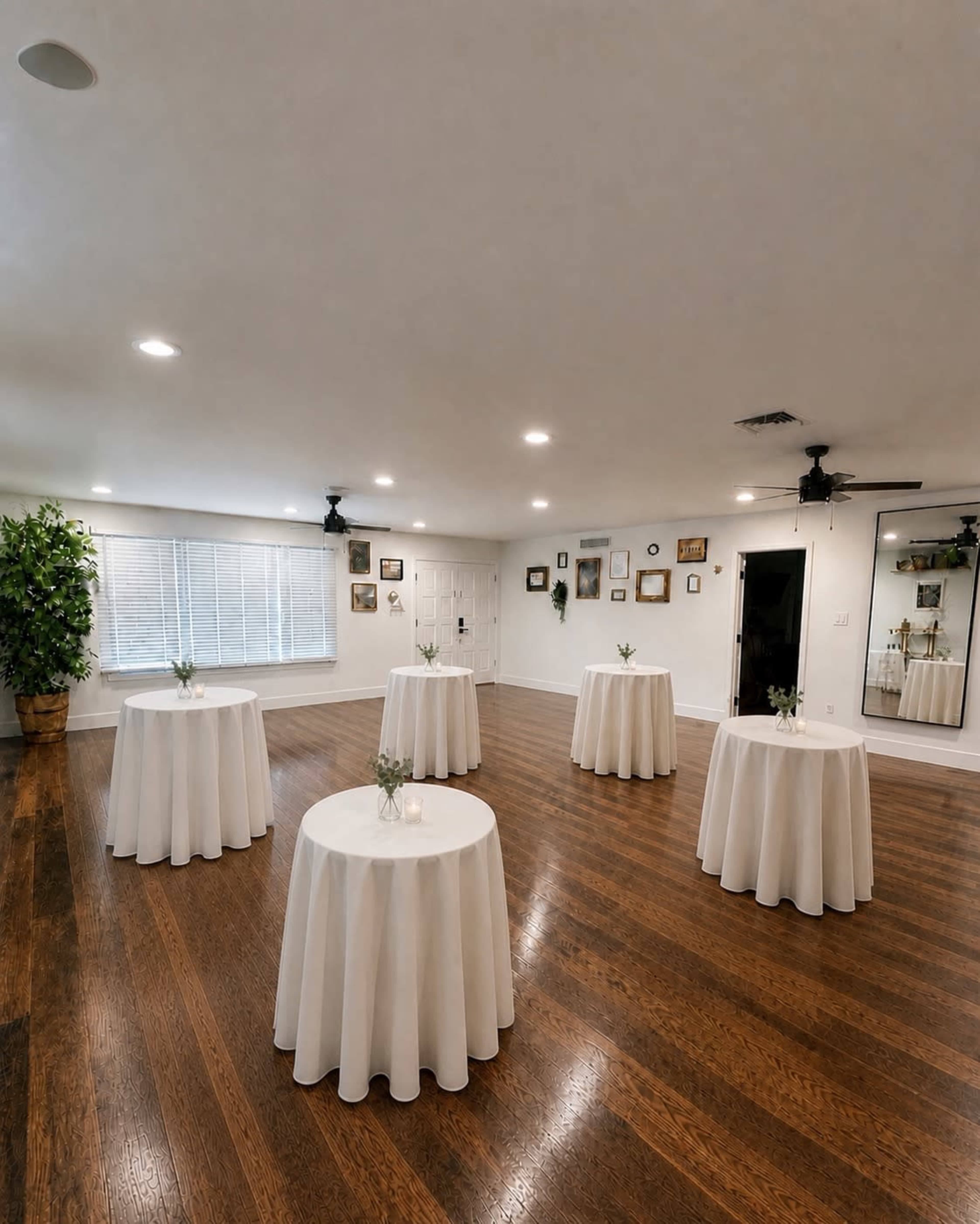 The image shows a spacious room with wooden flooring, featuring several round tables covered with white tablecloths and small floral centerpieces.