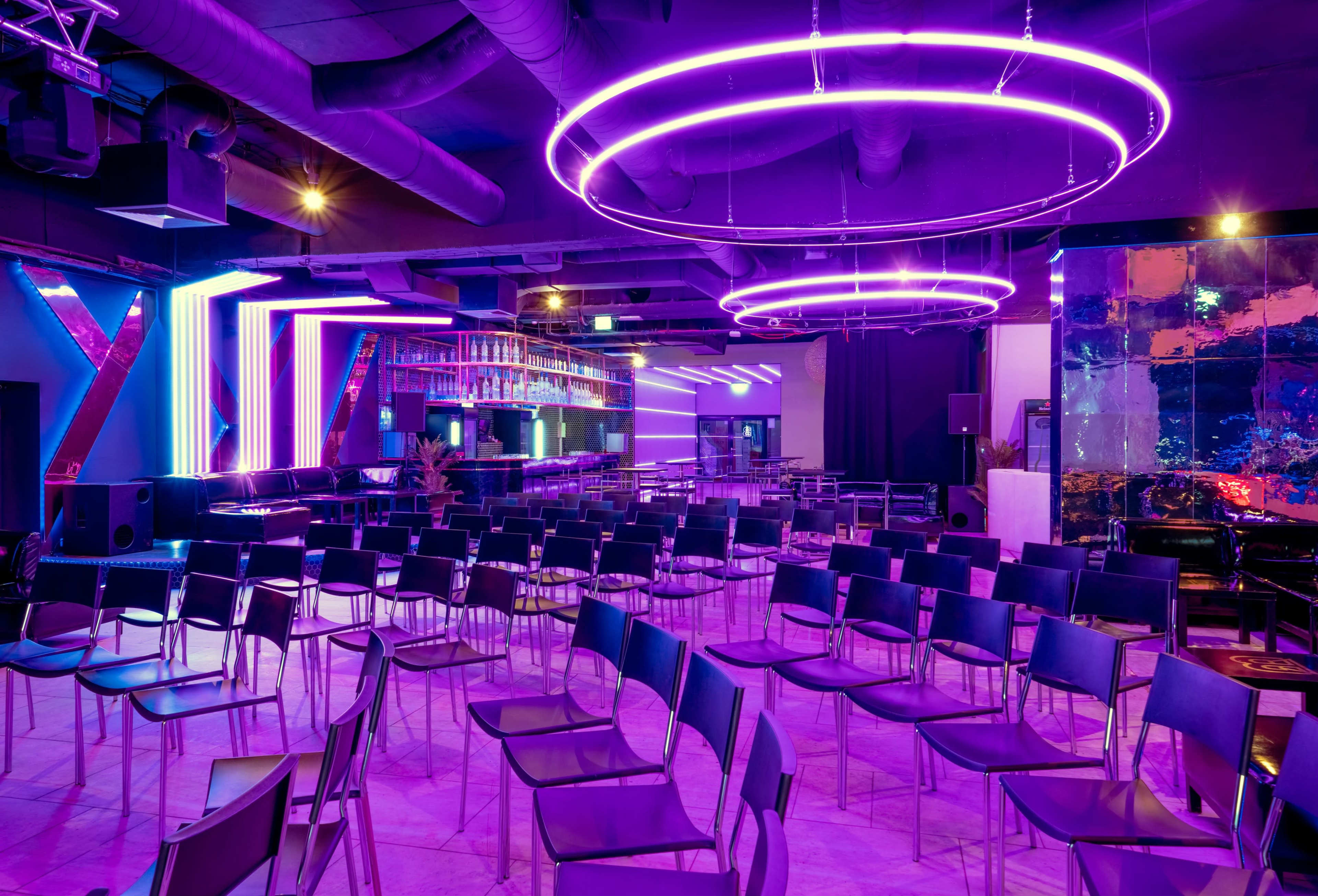 A modern setting with rows of black chairs arranged in front of a bar area, featuring colorful neon lights and a stage backdrop.