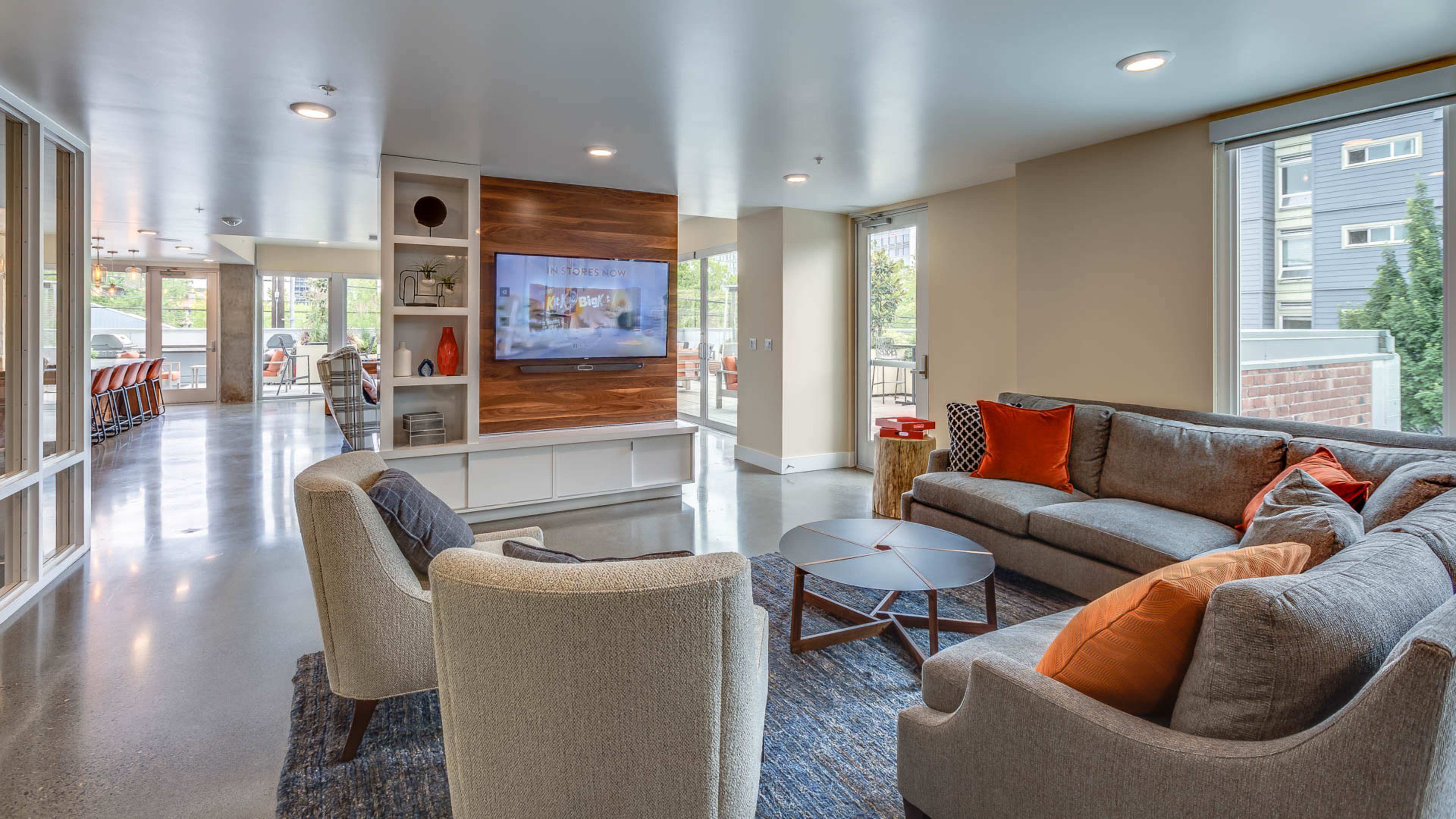 The image shows a modern living area with a gray sectional sofa, armchairs, a round coffee table, and a large TV mounted on a wooden feature wall.