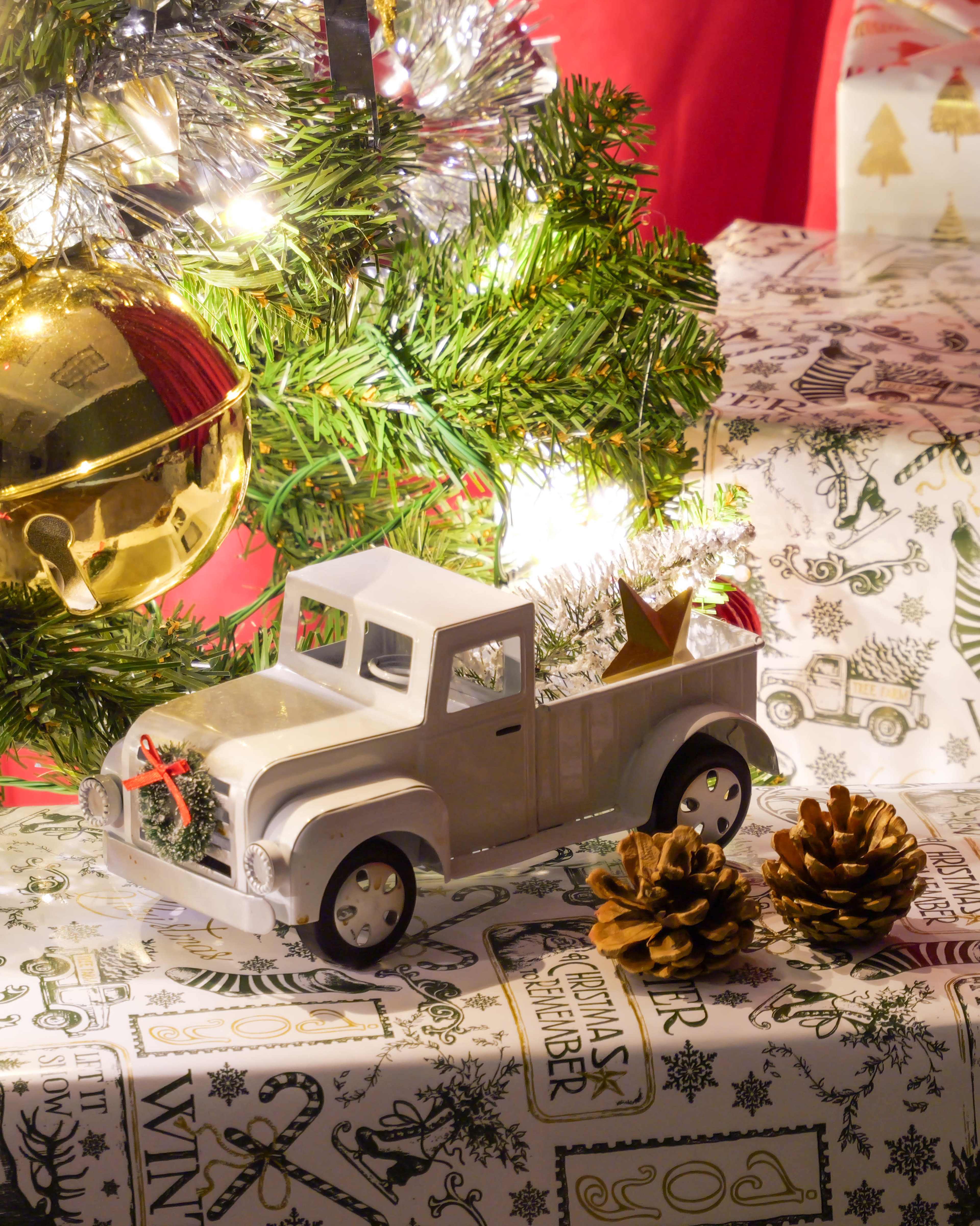 A white toy truck adorned with holiday decorations sits beside pine cones under a Christmas tree.