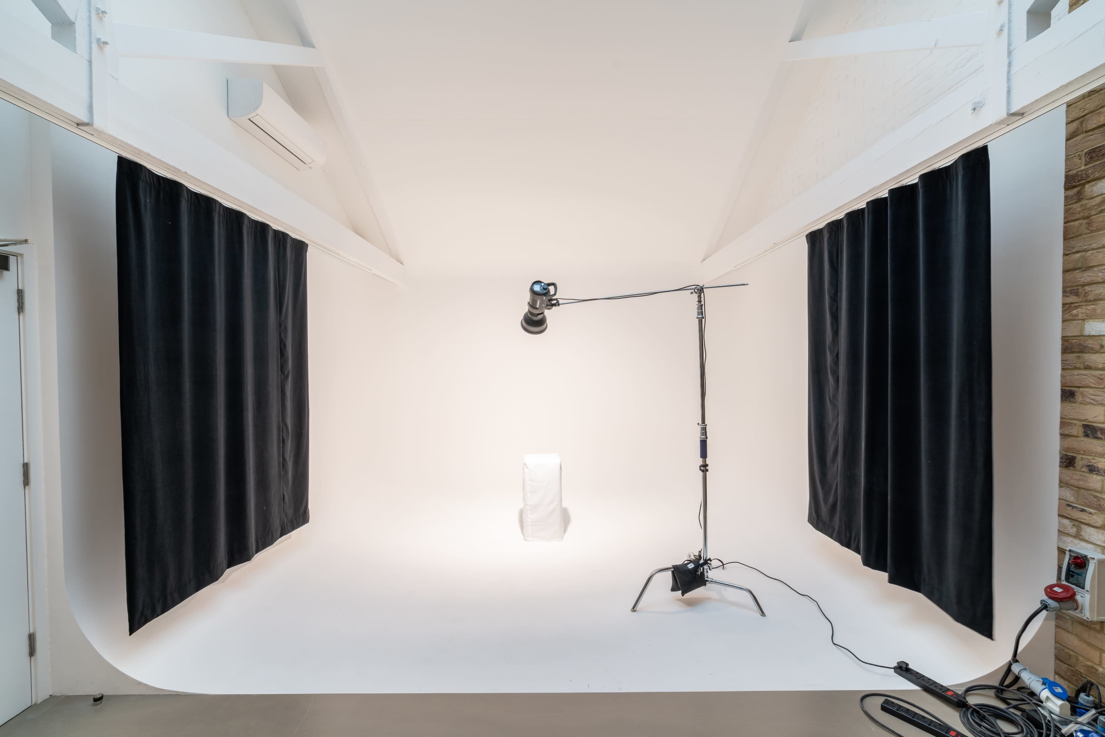 A photography studio with white walls, dark curtains on two sides, and a light stand positioned over a blank pedestal.