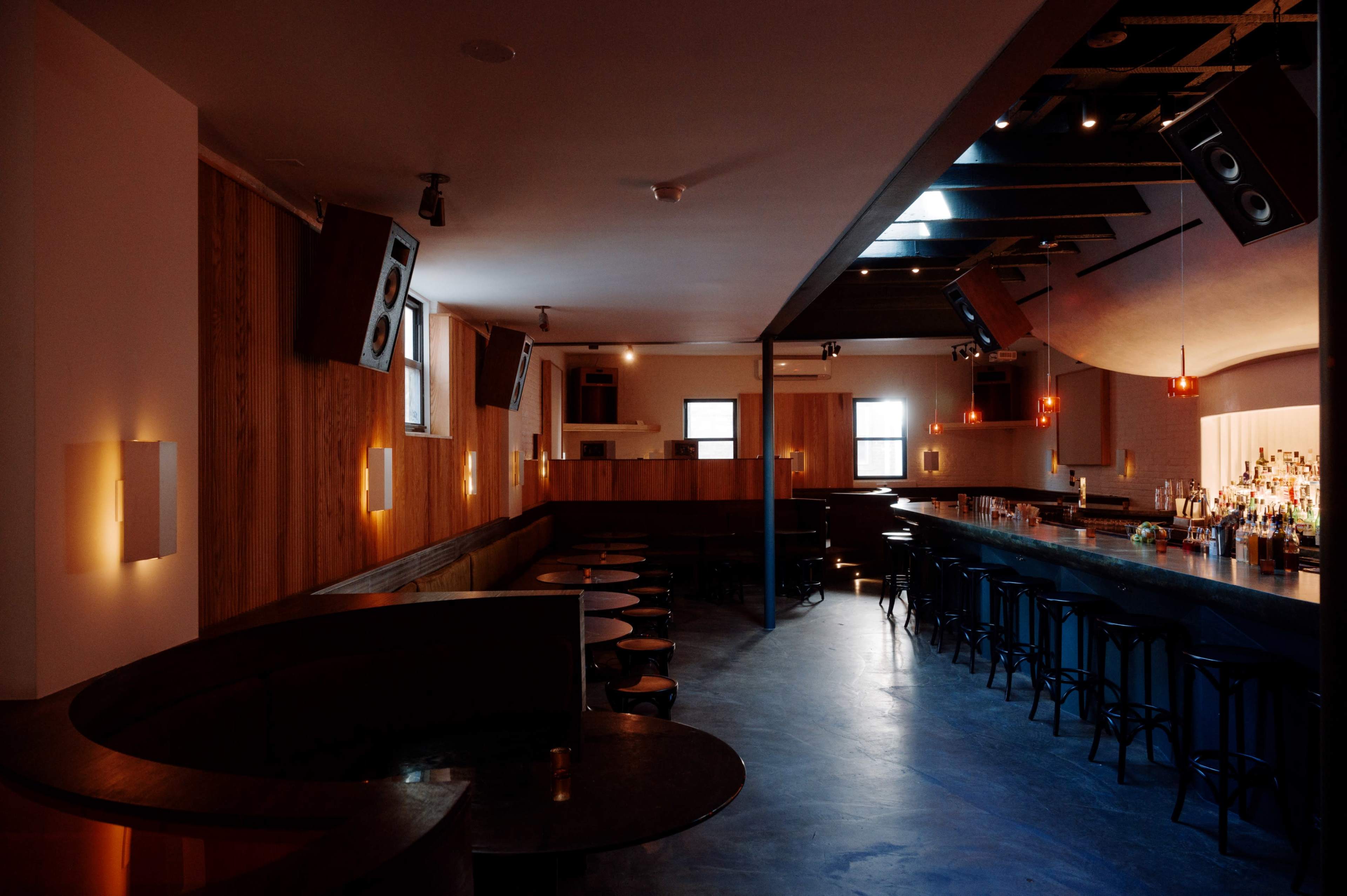 The image shows a dimly lit bar interior with wooden walls, a long counter stocked with alcohol, and scattered tables arranged along the floor.