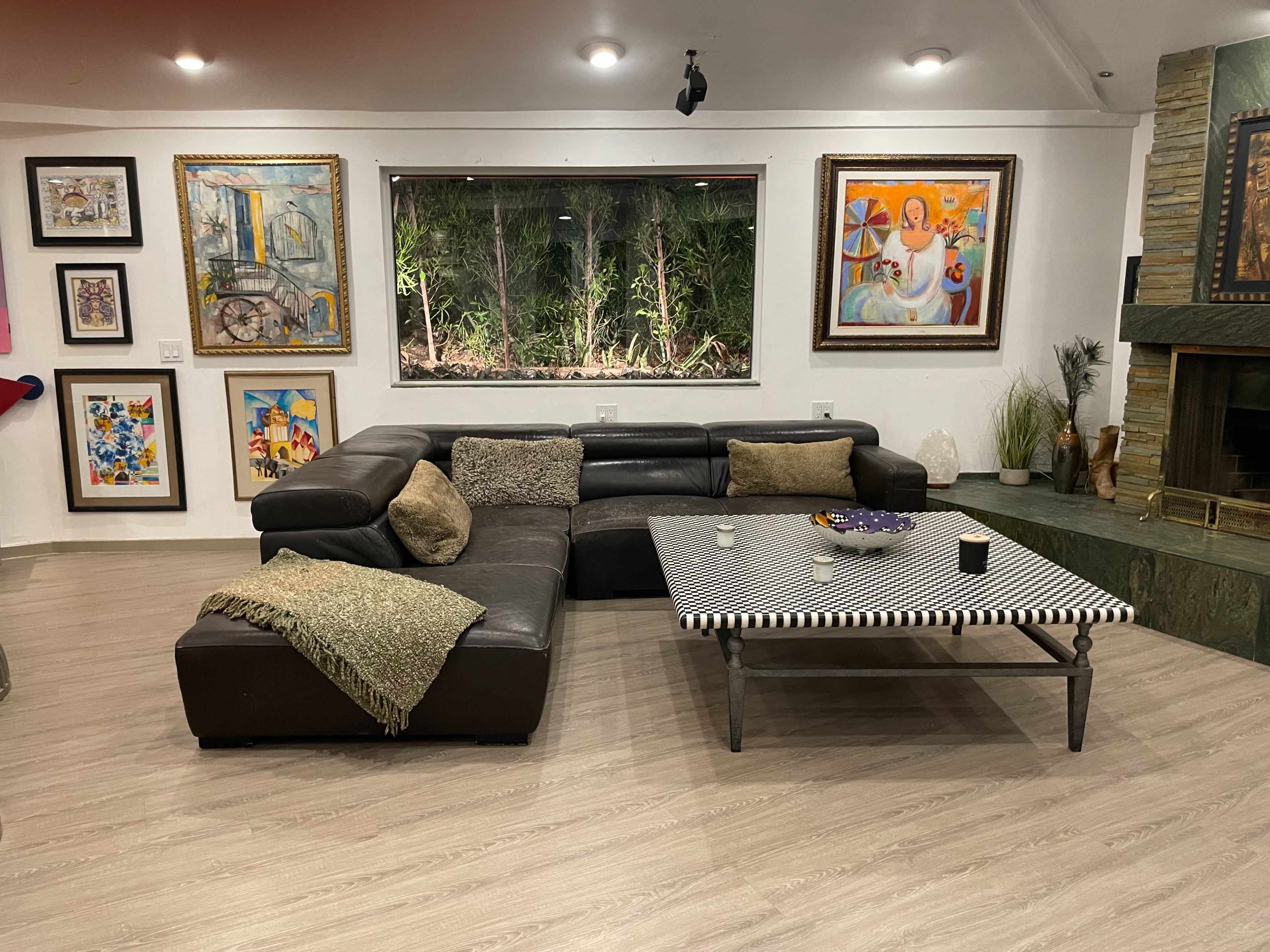 A living room with a large black sectional sofa, a striped coffee table, framed artwork on the walls, and a window overlooking greenery.