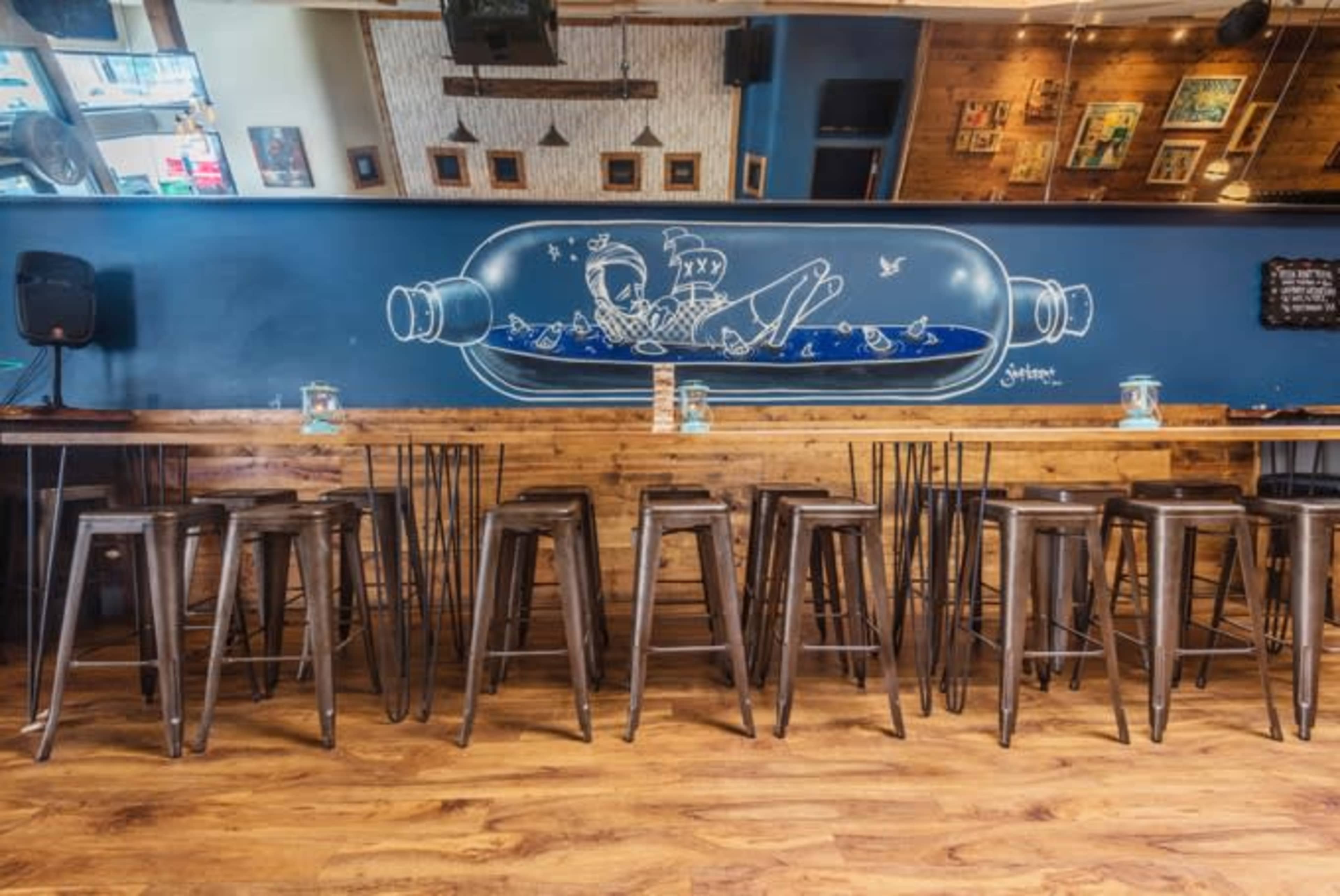 A modern bar with metal stools arranged in front of a wooden counter, adorned with a large mural of a bottle and a figure inside it on the blue wall behind.