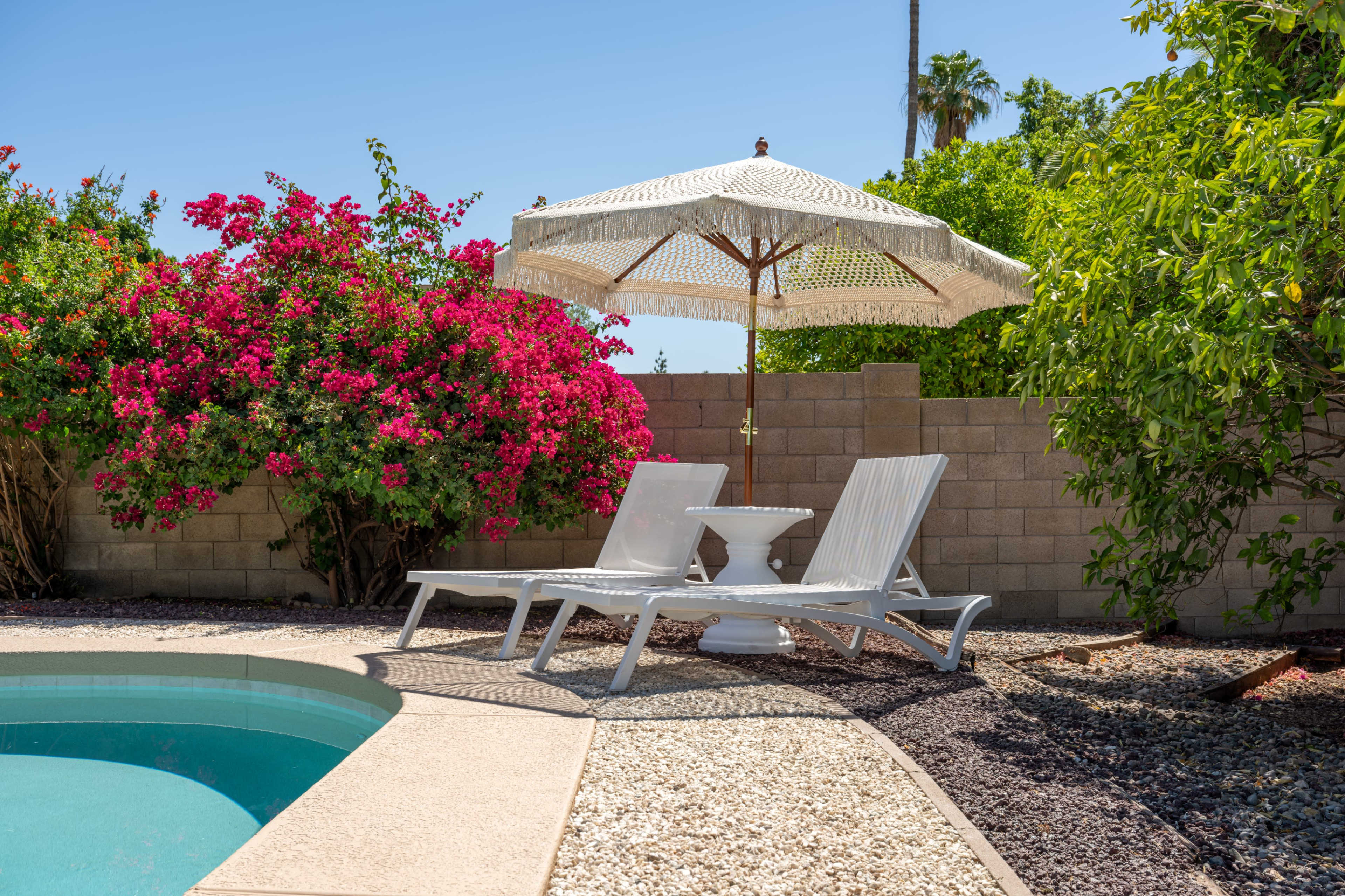 A patio area features two white lounge chairs and a decorative umbrella beside a swimming pool, surrounded by flowering plants and a stone wall.