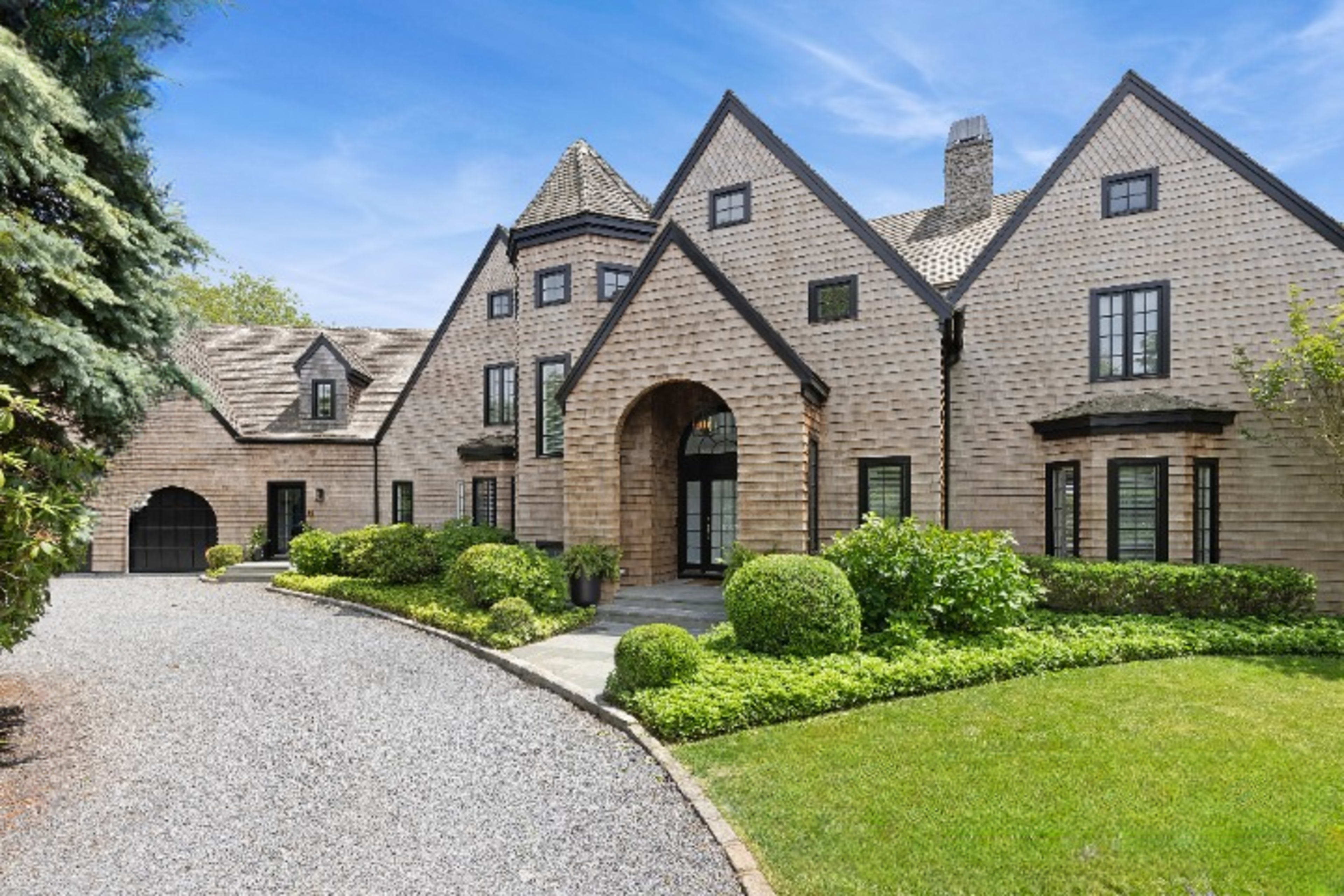 A large, multi-gabled house with a stone entrance and landscaped grounds is set along a gravel driveway.
