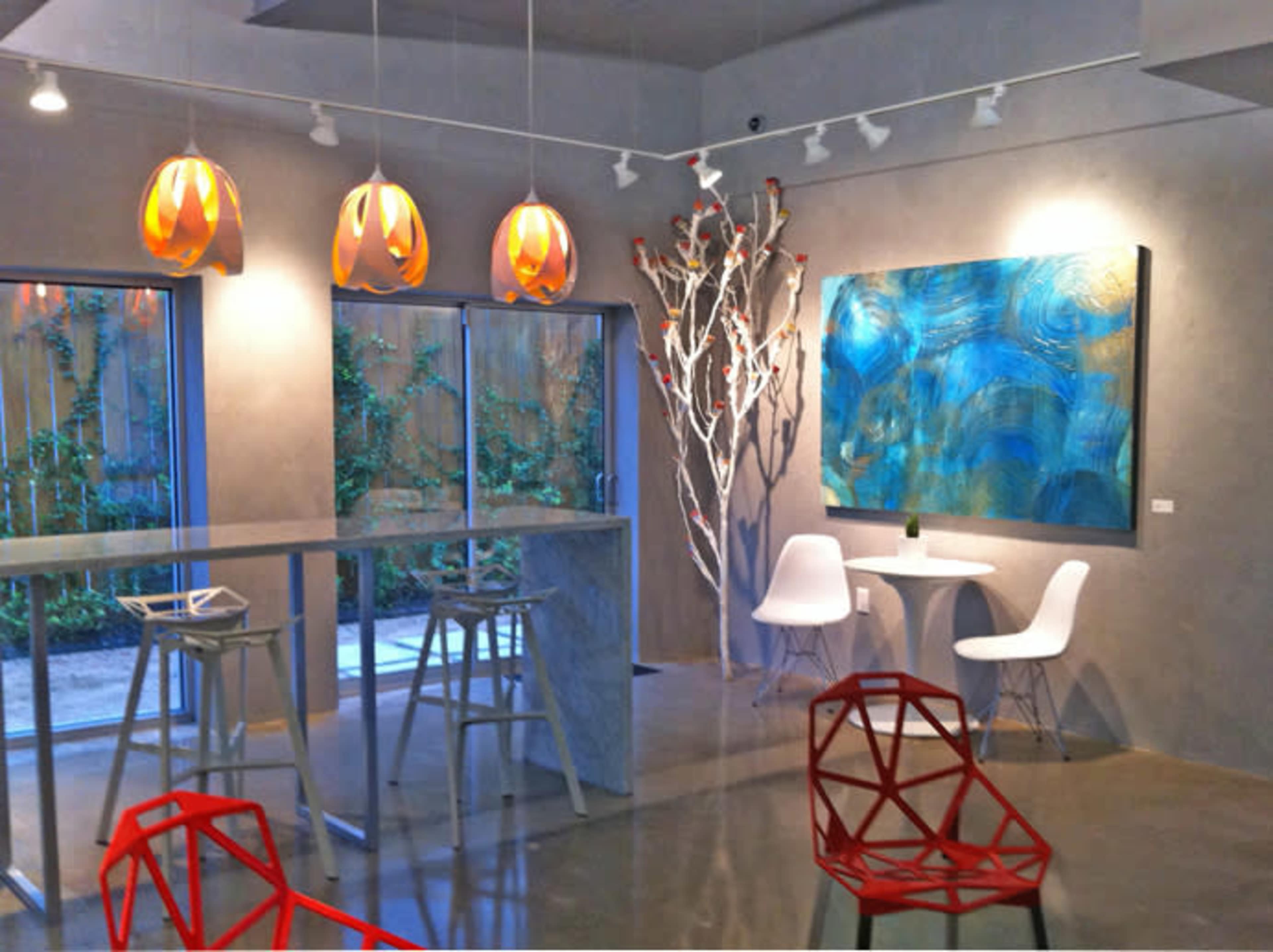 The image shows a modern, minimalistic art gallery featuring a large blue painting on the wall, display tables, unique light fixtures, and decorative tree-like sculptures.