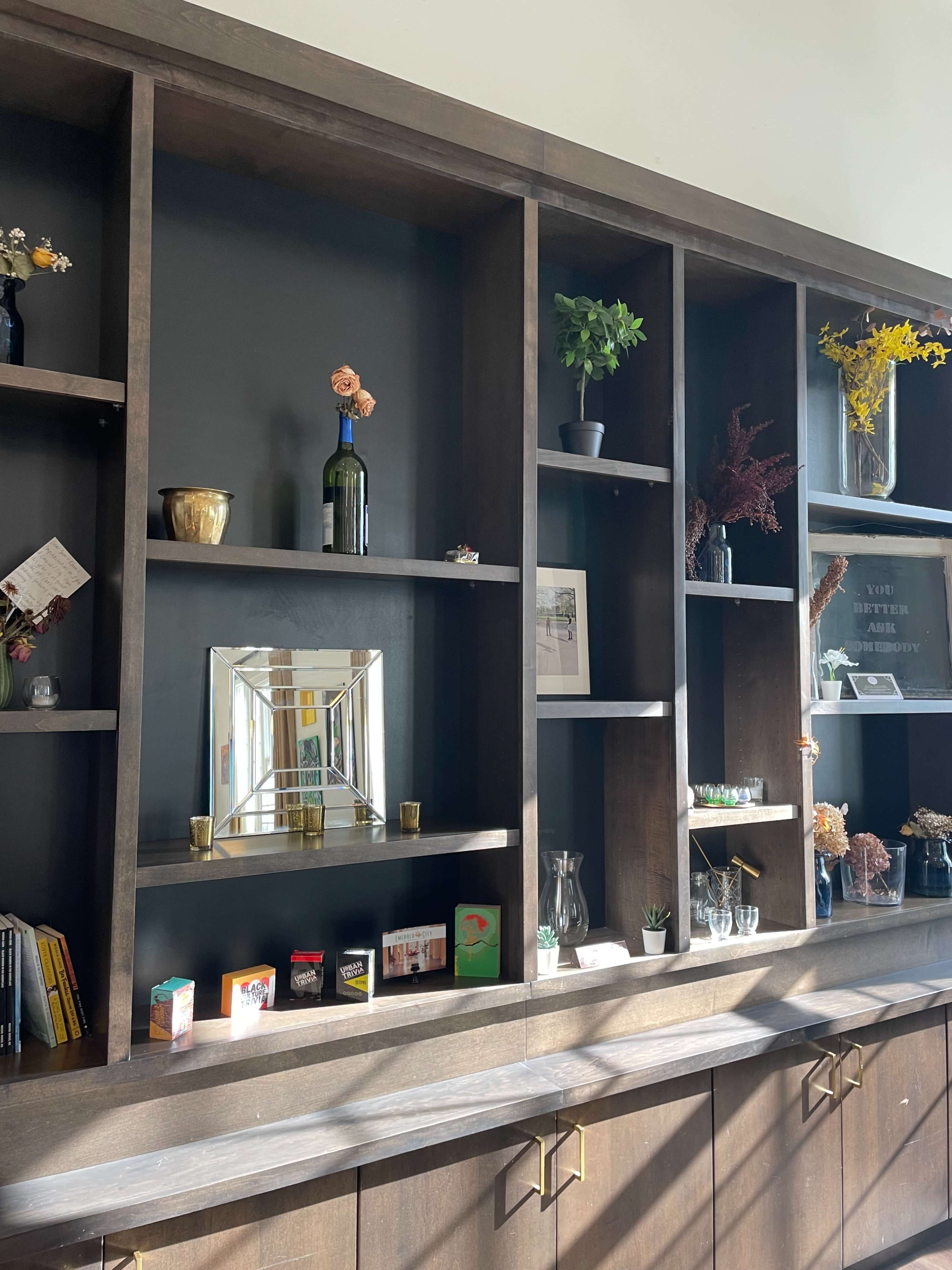 A wooden shelving unit against a dark wall, decorated with various plants, vases, and framed items, with several small objects displayed on the shelves.