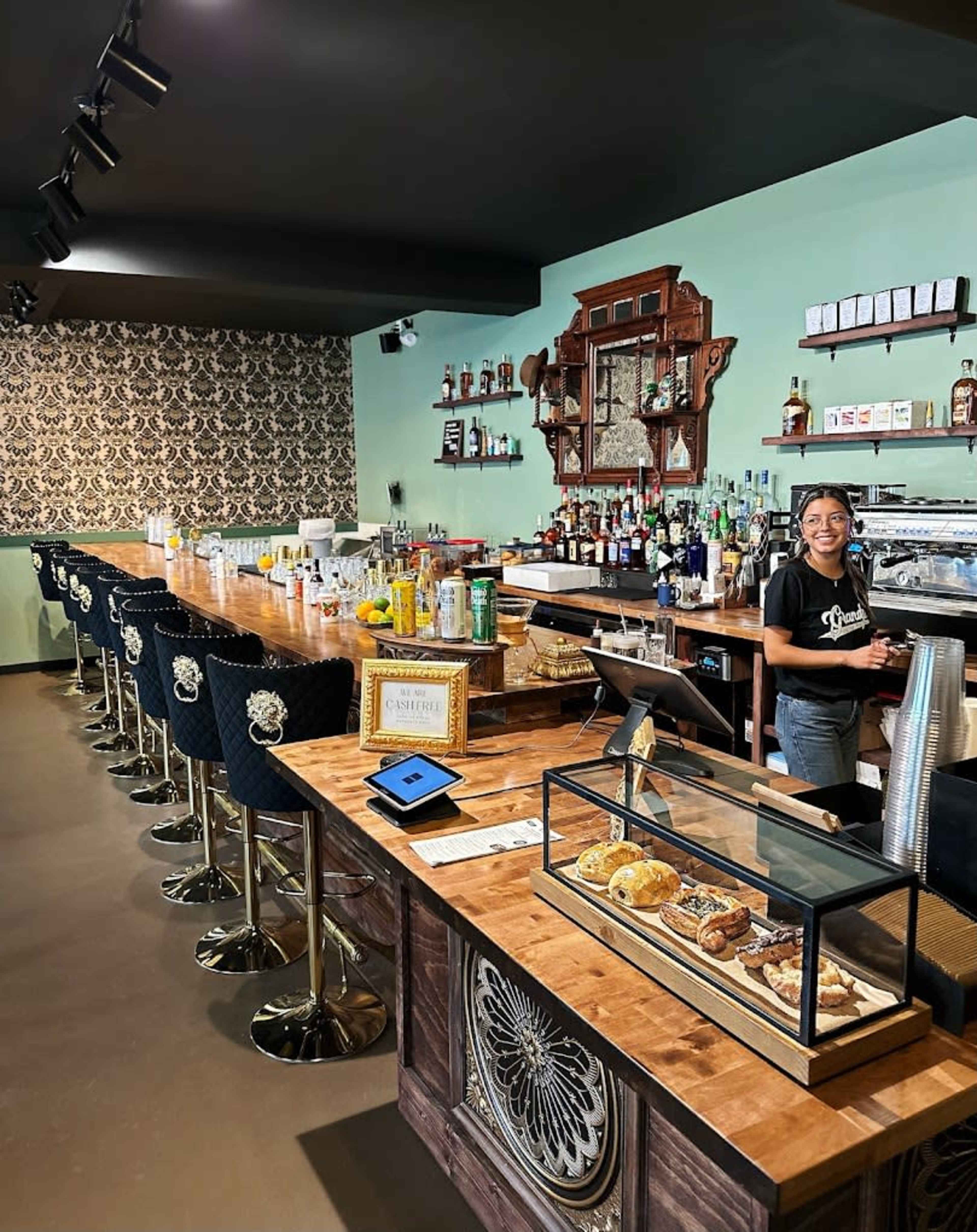 The image shows a stylish bar with a wooden counter, high stools, a variety of liquor bottles on shelves, and a display case of pastries, along with a staff member standing at the bar.