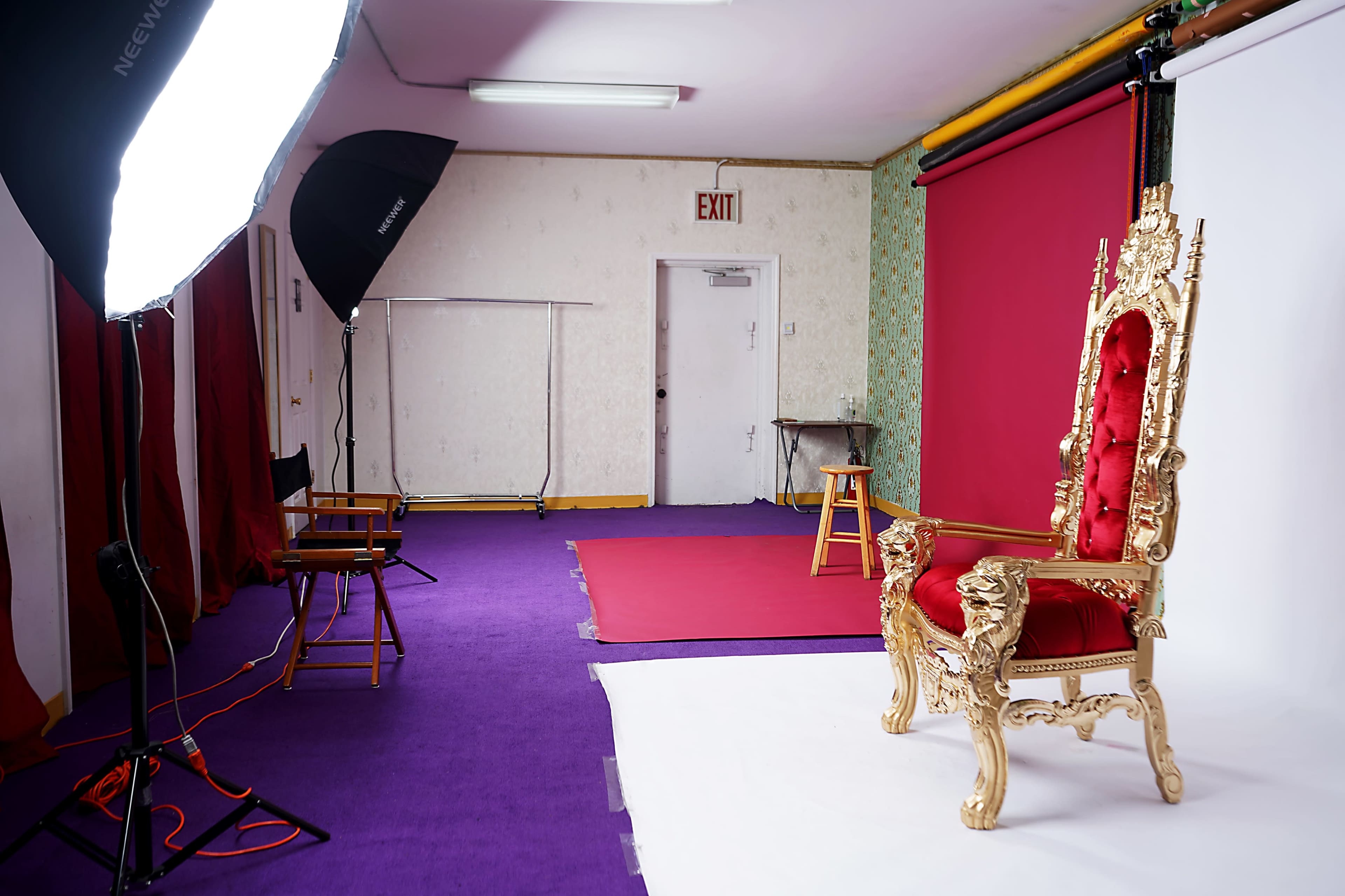 A photography studio with a gold throne chair on a white backdrop, surrounded by lighting equipment and a purple carpet.