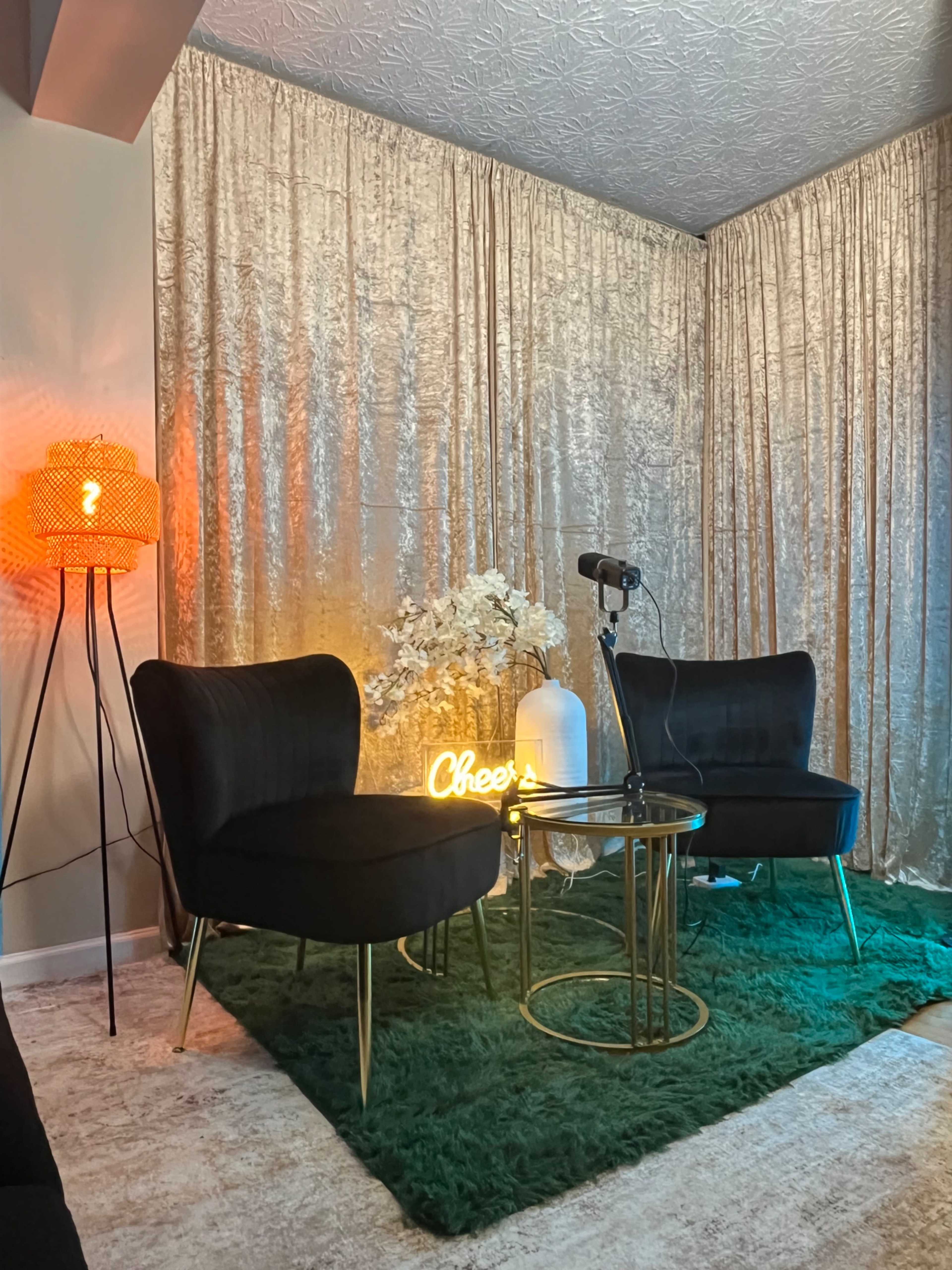 The image shows a cozy seating area with two black chairs, a round coffee table, decorative flowers, and an illuminated sign against a backdrop of golden curtains.