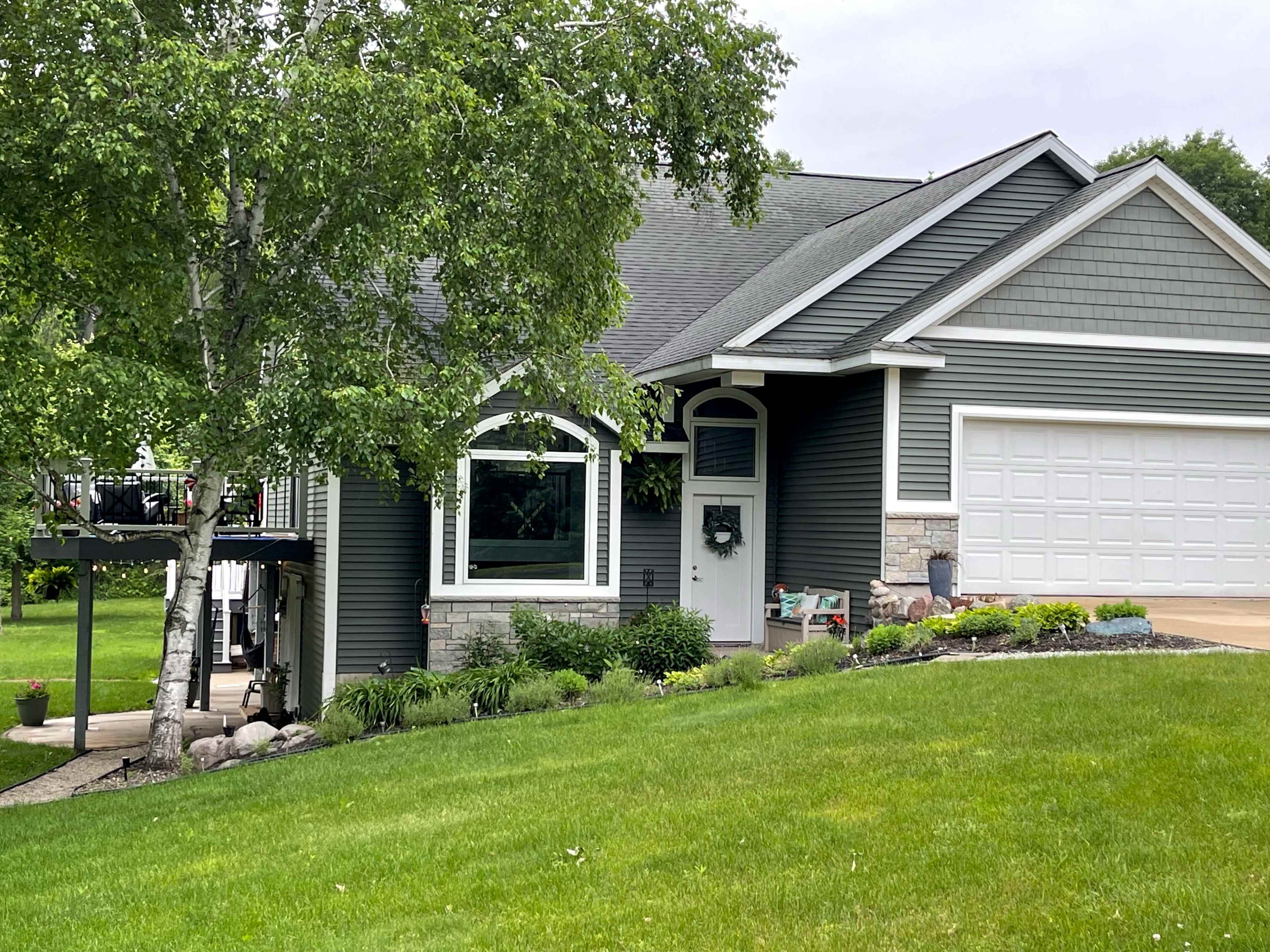 A two-story house with gray siding and a landscaped front yard is shown, featuring a driveway and a large garage door.
