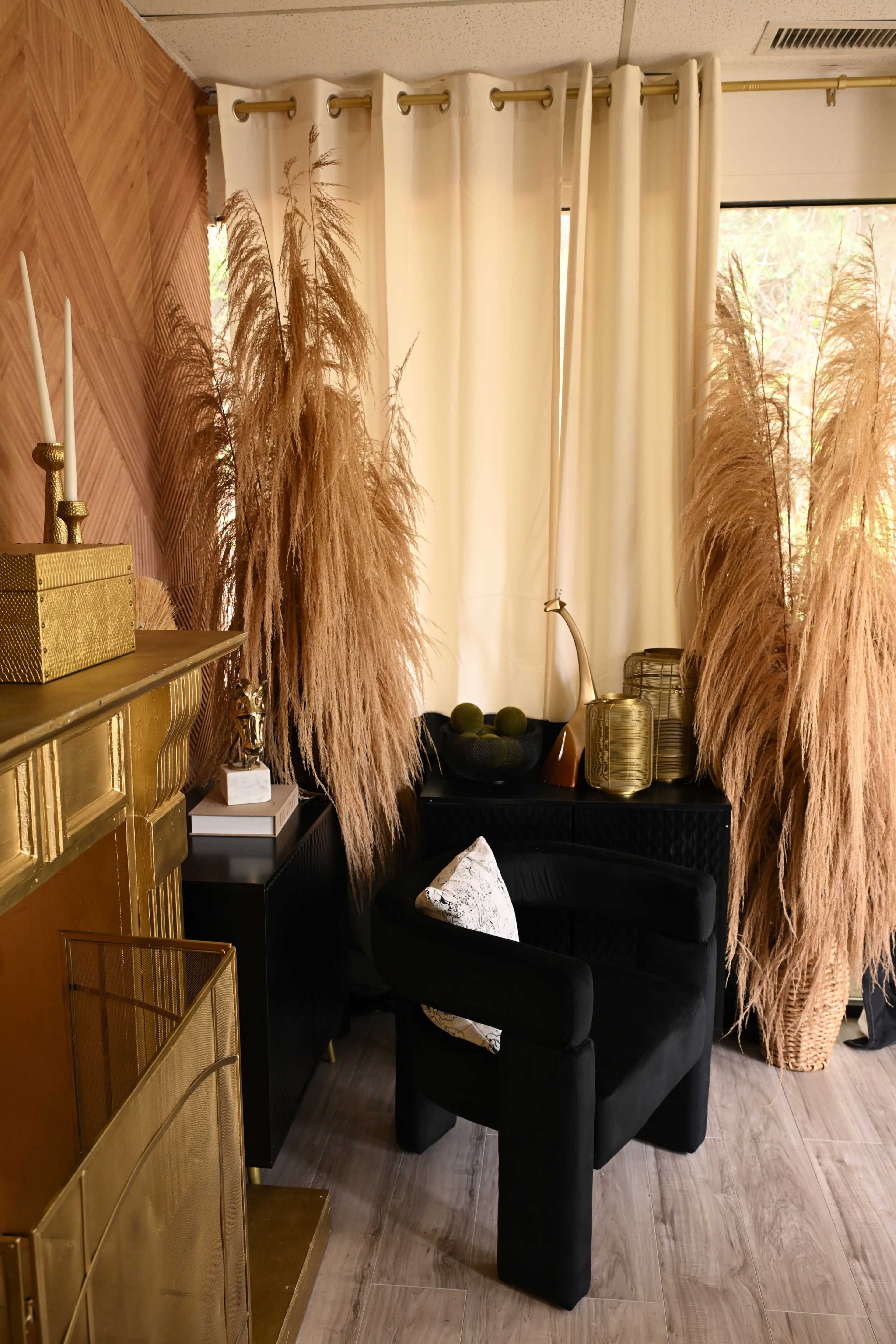 The image shows a cozy corner of a room featuring a black armchair, a fireplace with a gold surround, and tall, decorative dried plants beside large windows covered by light curtains.