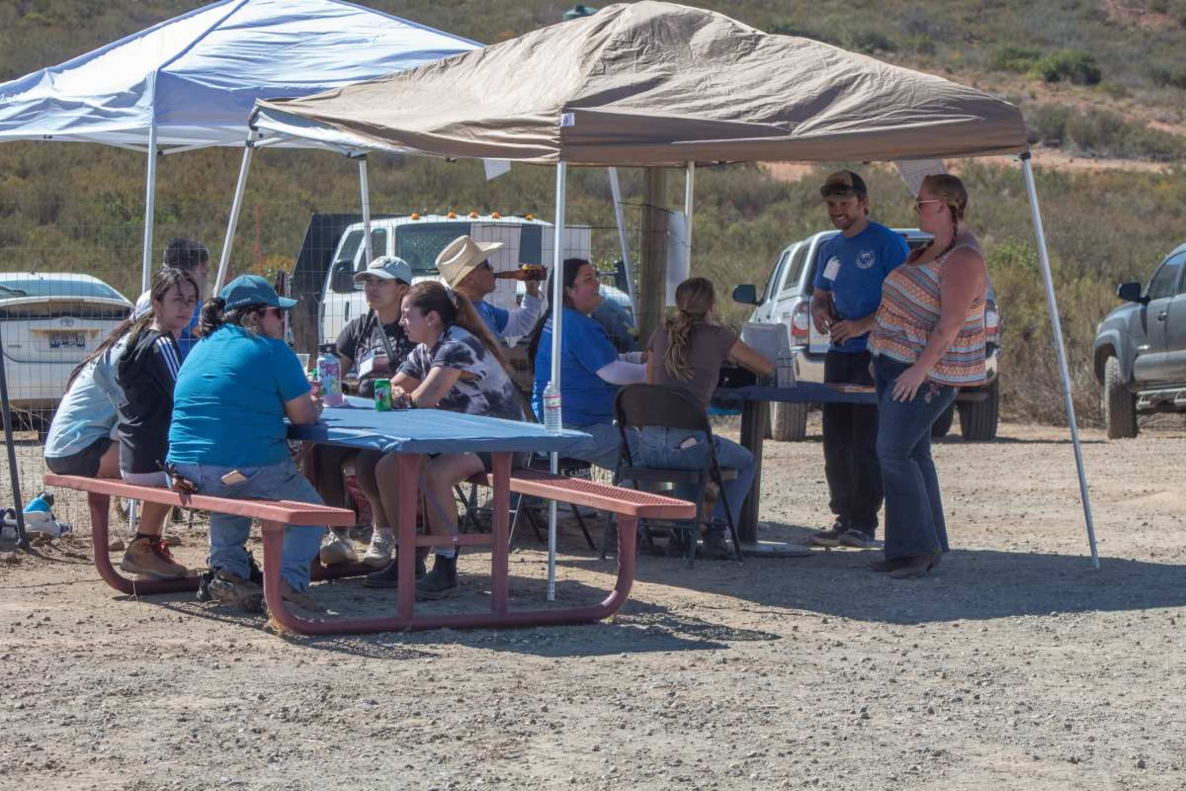 A group of people is gathered around picnic tables under tents in an outdoor setting, with vehicles parked nearby.