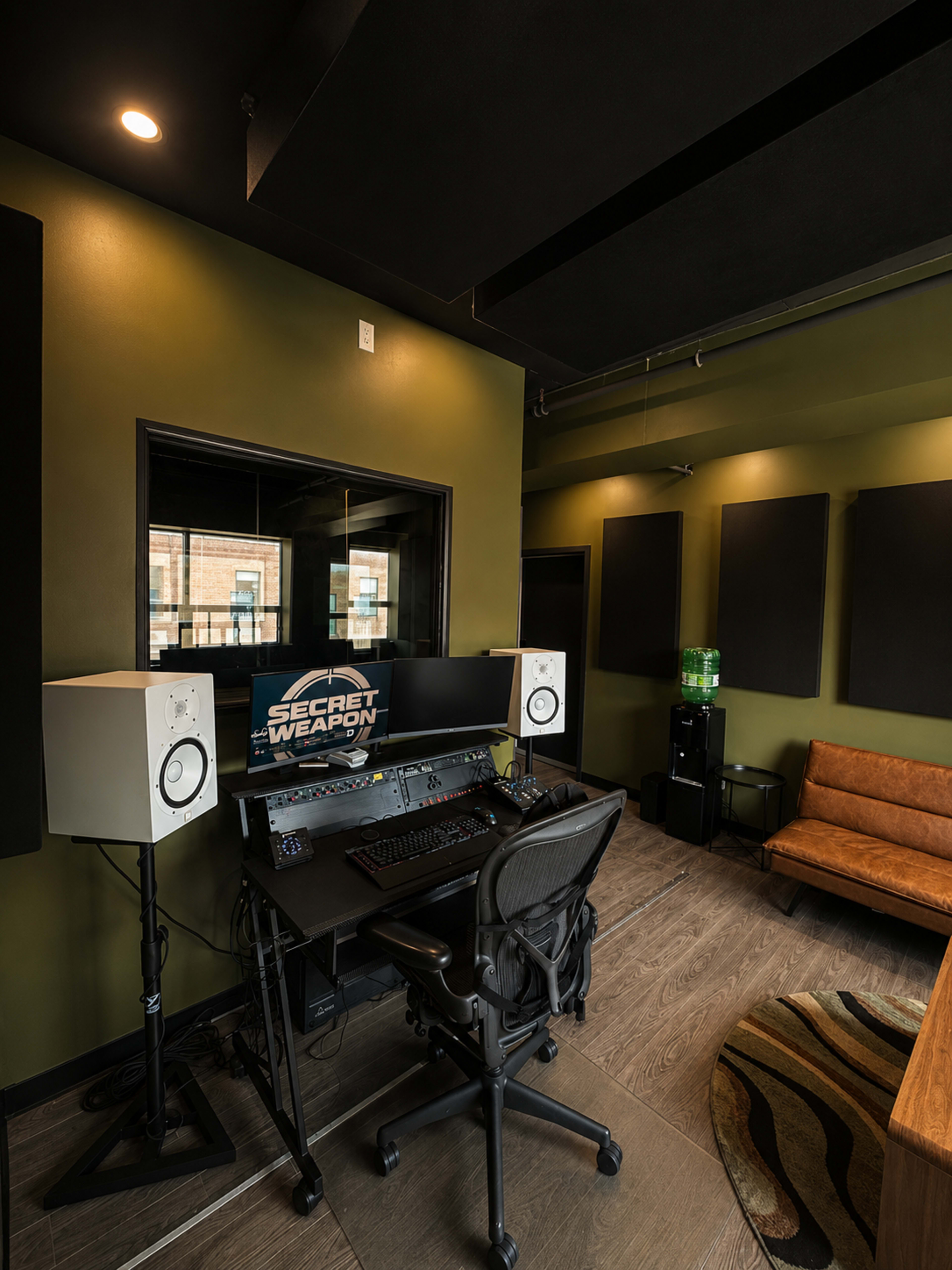 The image shows a recording studio with a desk featuring audio equipment, two speaker monitors, and a brown couch against a green wall.