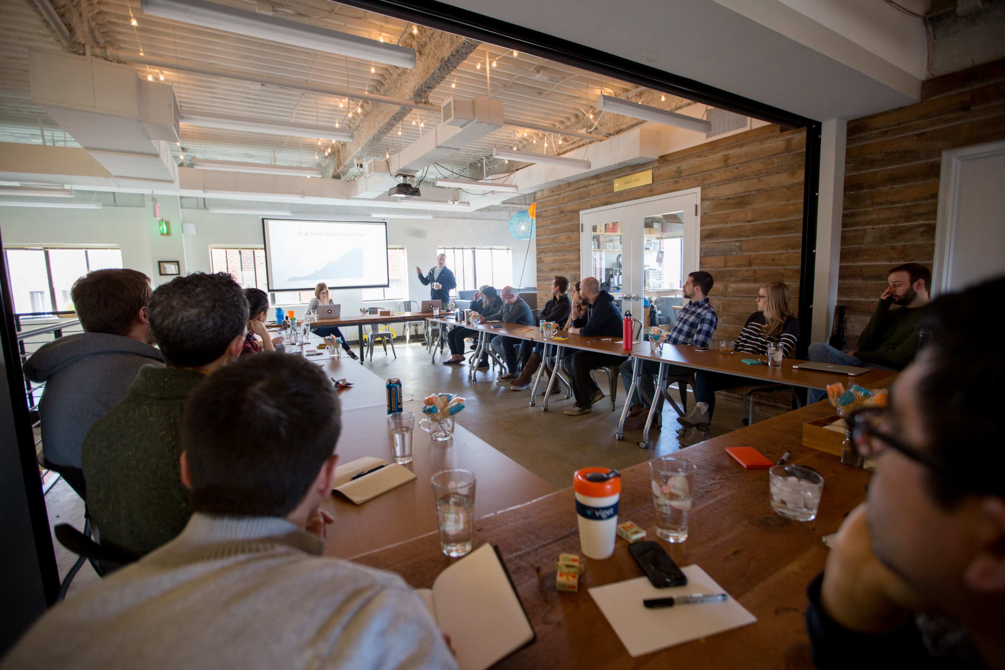 A group of people is seated around a large table in a meeting room, while one person stands at the front presenting to the audience.