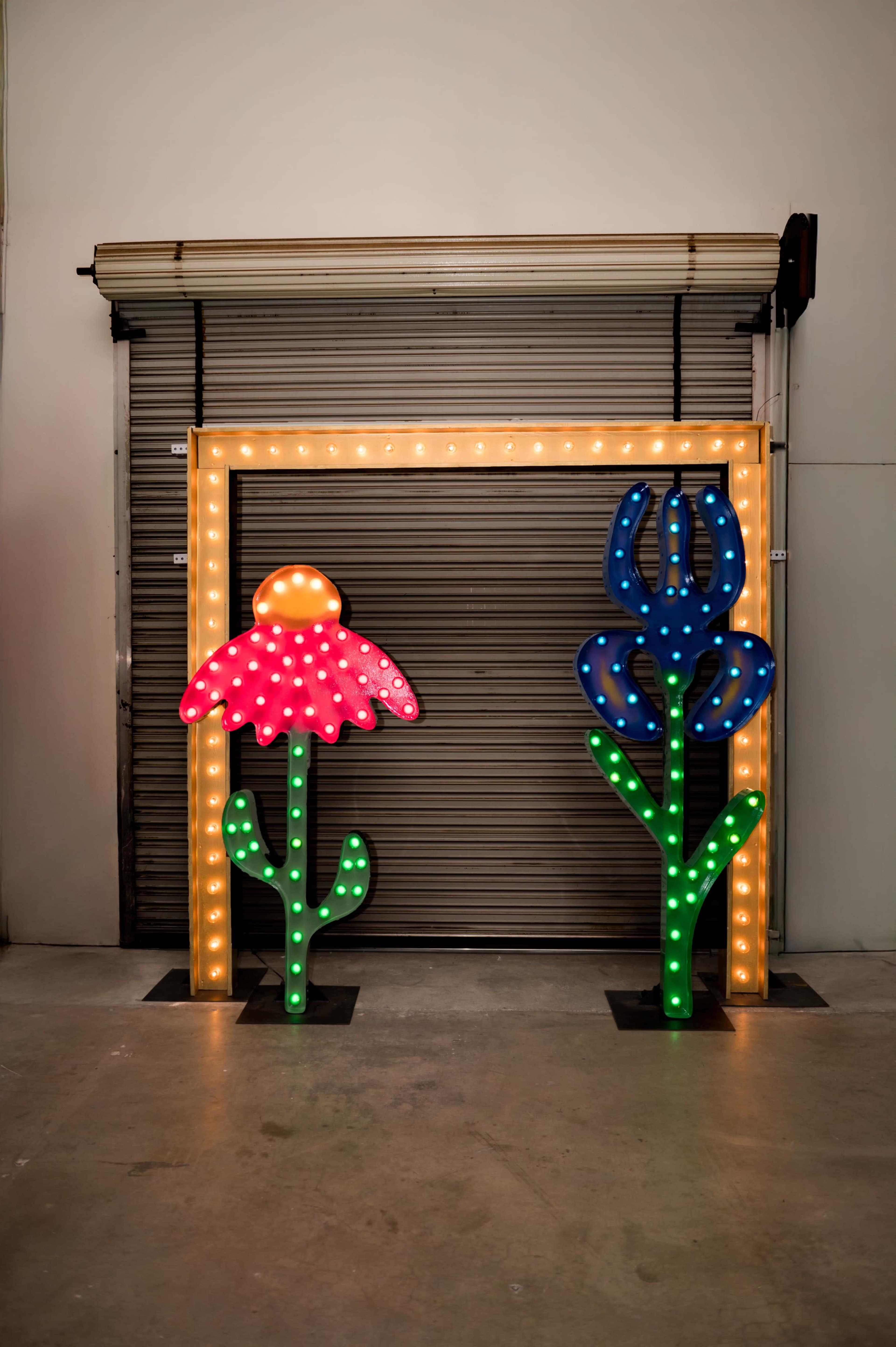 The image shows a brightly lit floral archway with two oversized flower sculptures, one red and one blue, on either side of a closed garage door.