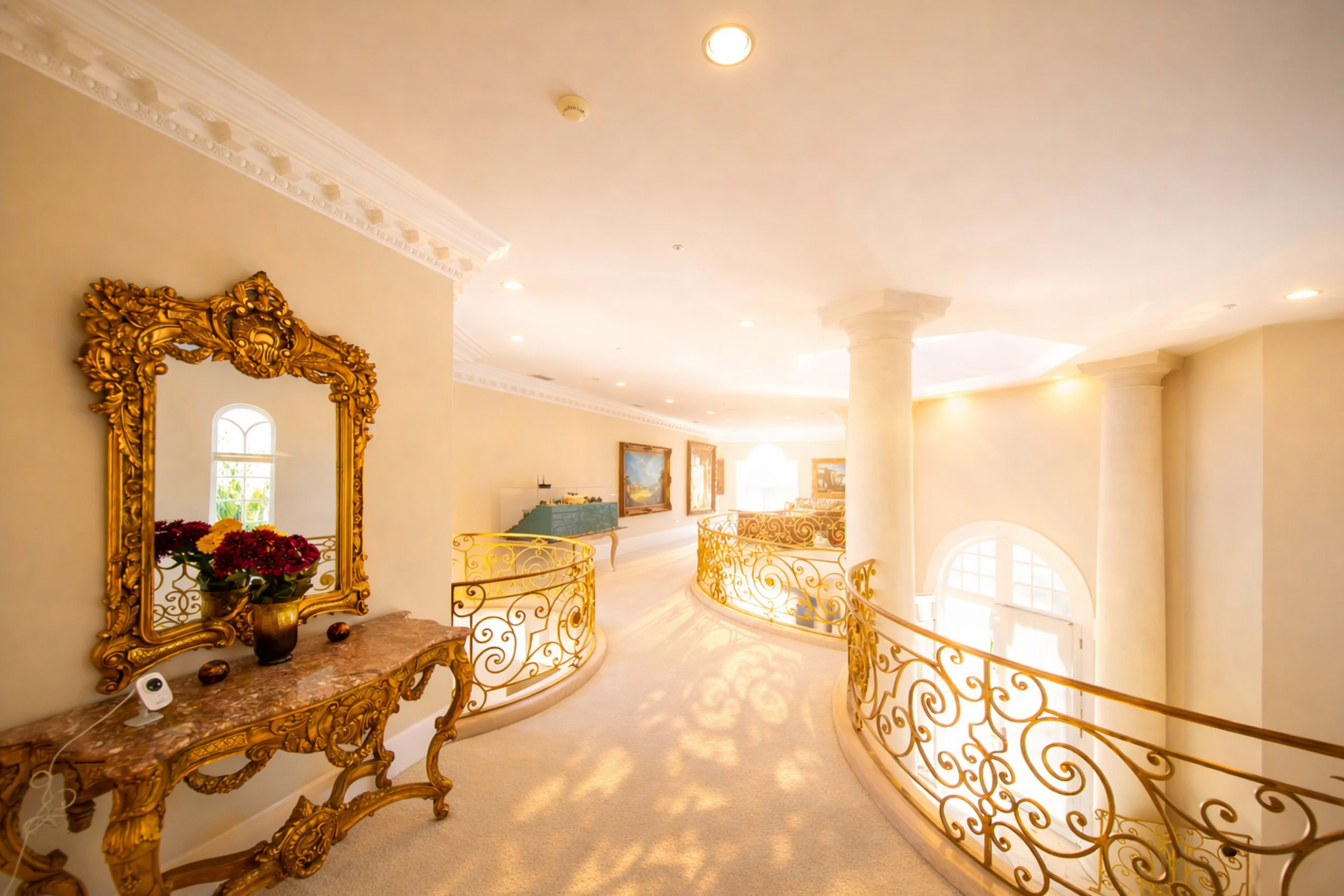 The image shows a brightly lit hallway with ornate gold railings, a decorative mirror, and paintings hung on the walls.