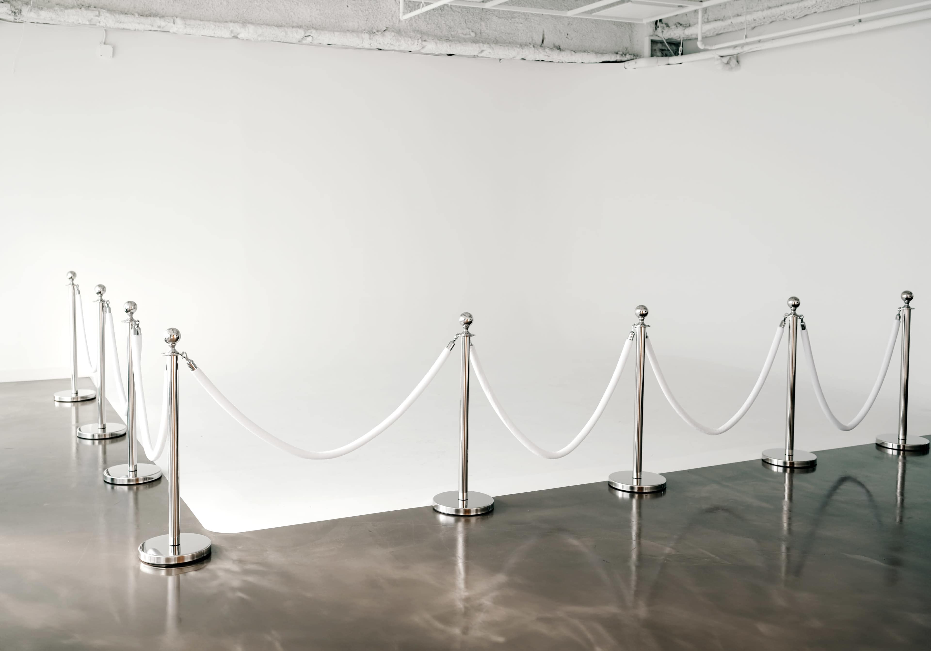A series of polished stanchions with white ropes arranged in a roped-off section of a minimalist, empty room.