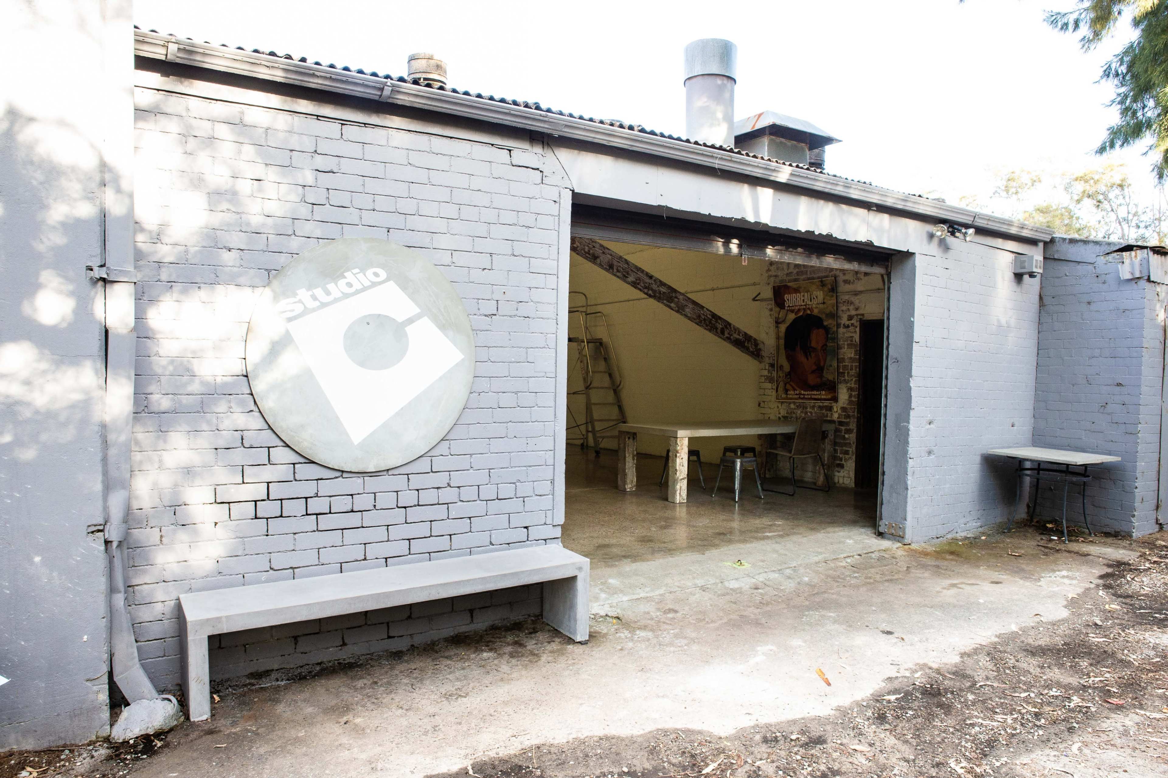 The image shows a gray brick building with an oval sign labeled "Studio" and an open entrance revealing a minimally furnished interior space.