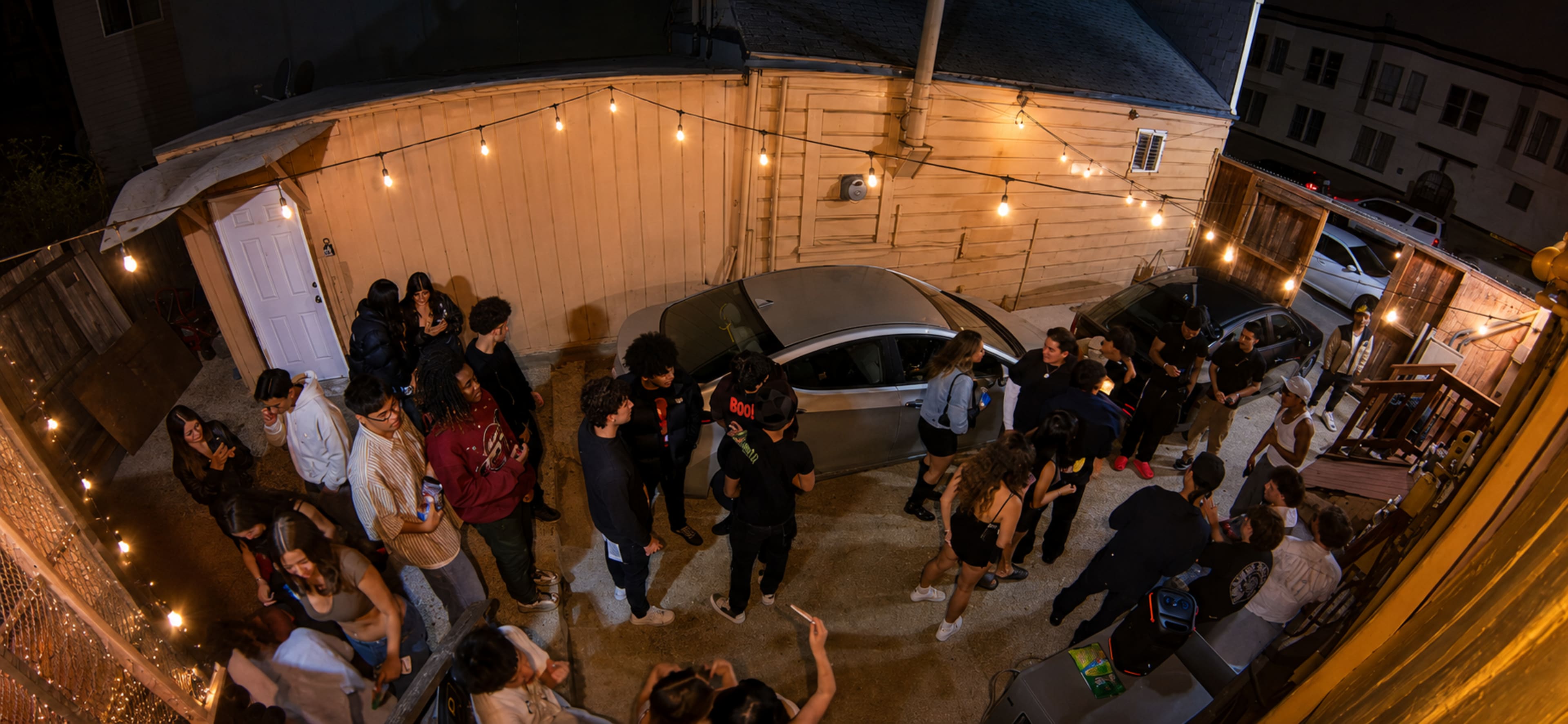 A group of people gathers in a dimly lit outdoor space adorned with string lights, surrounded by parked cars and a building exterior.