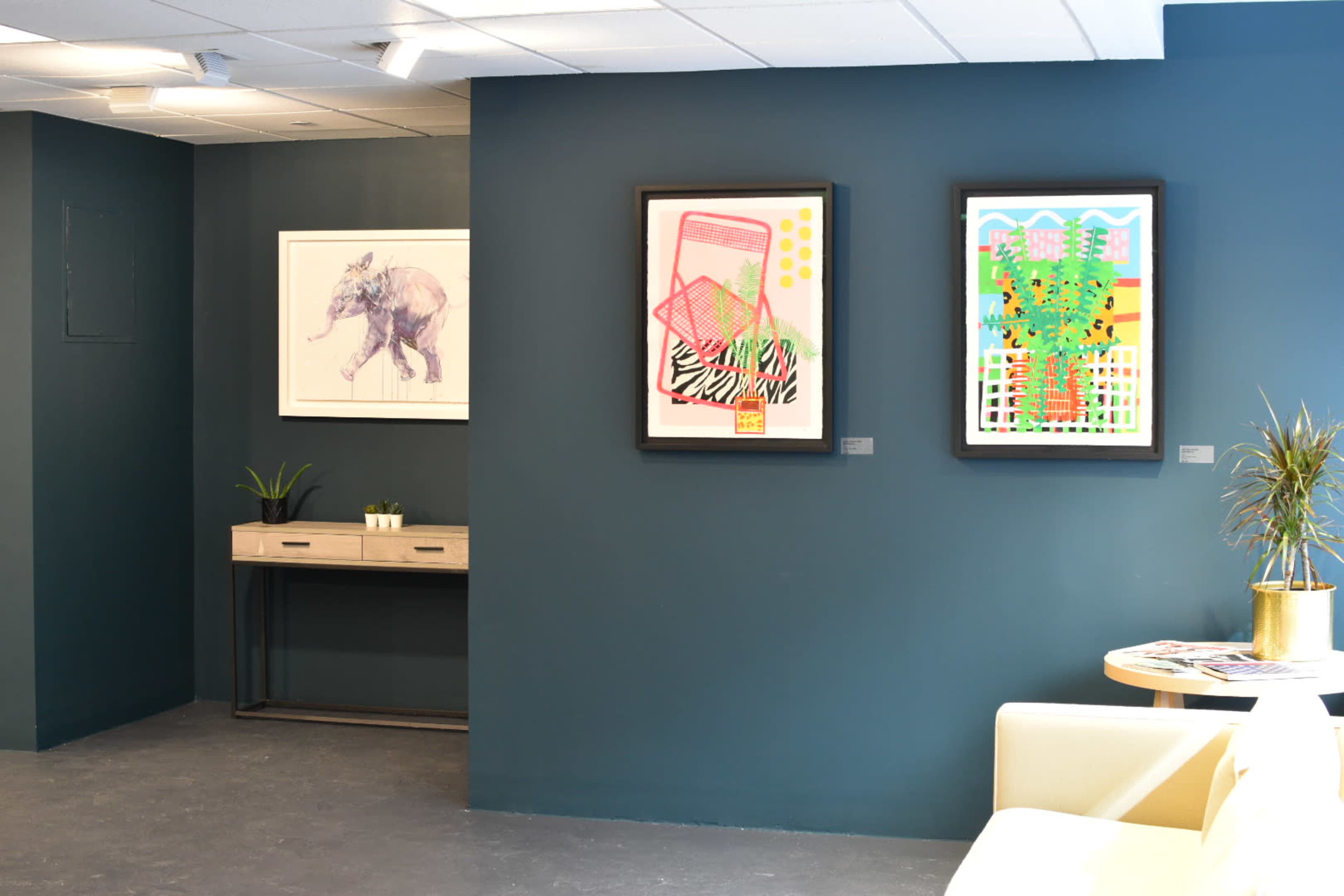 The image shows an interior wall with two framed artworks and a small console table adorned with a plant.