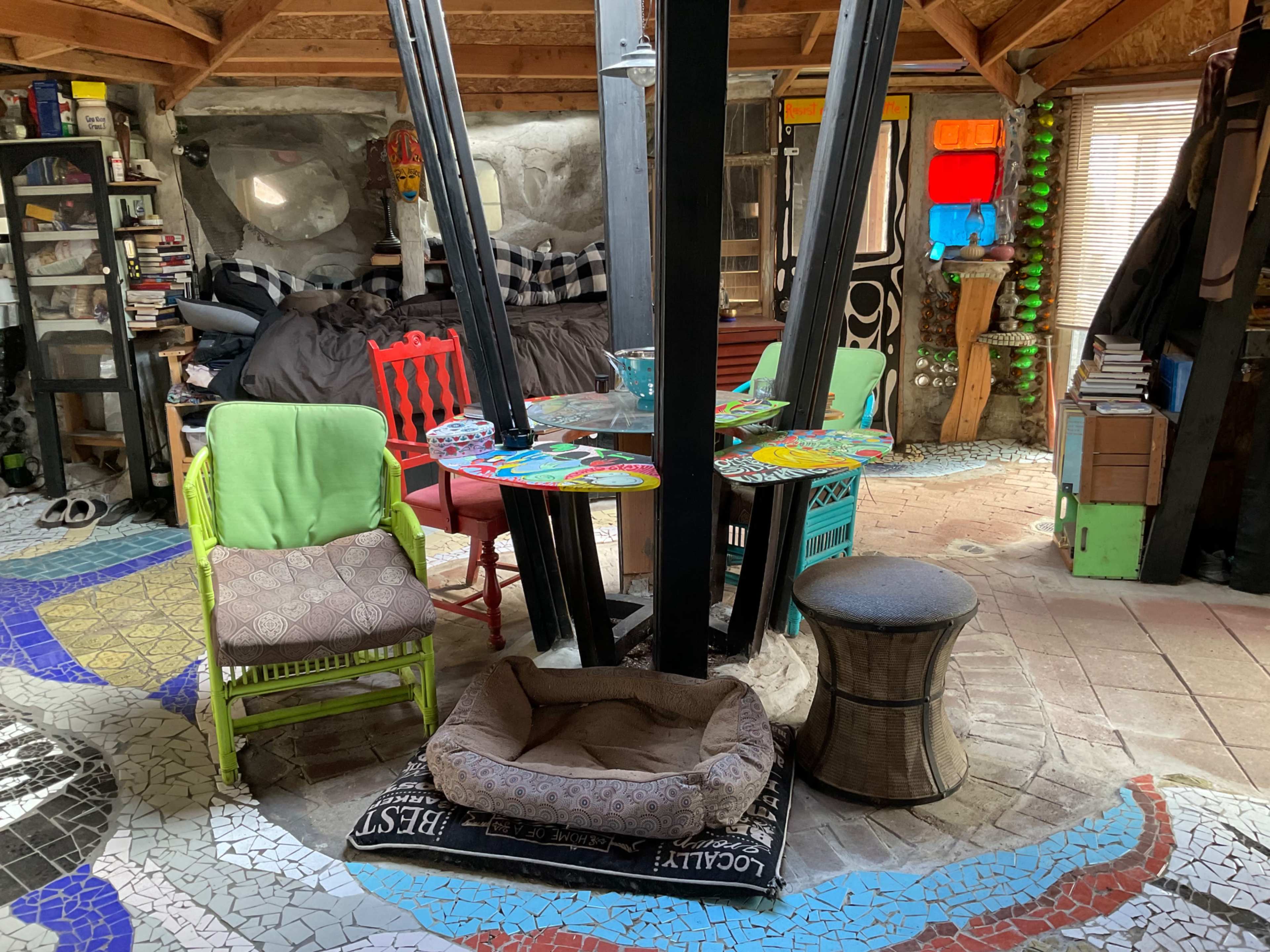 The image shows an eclectic room featuring colorful chairs surrounding a table with a mosaic floor, a bed in the background, and various shelves and decorations on the walls.