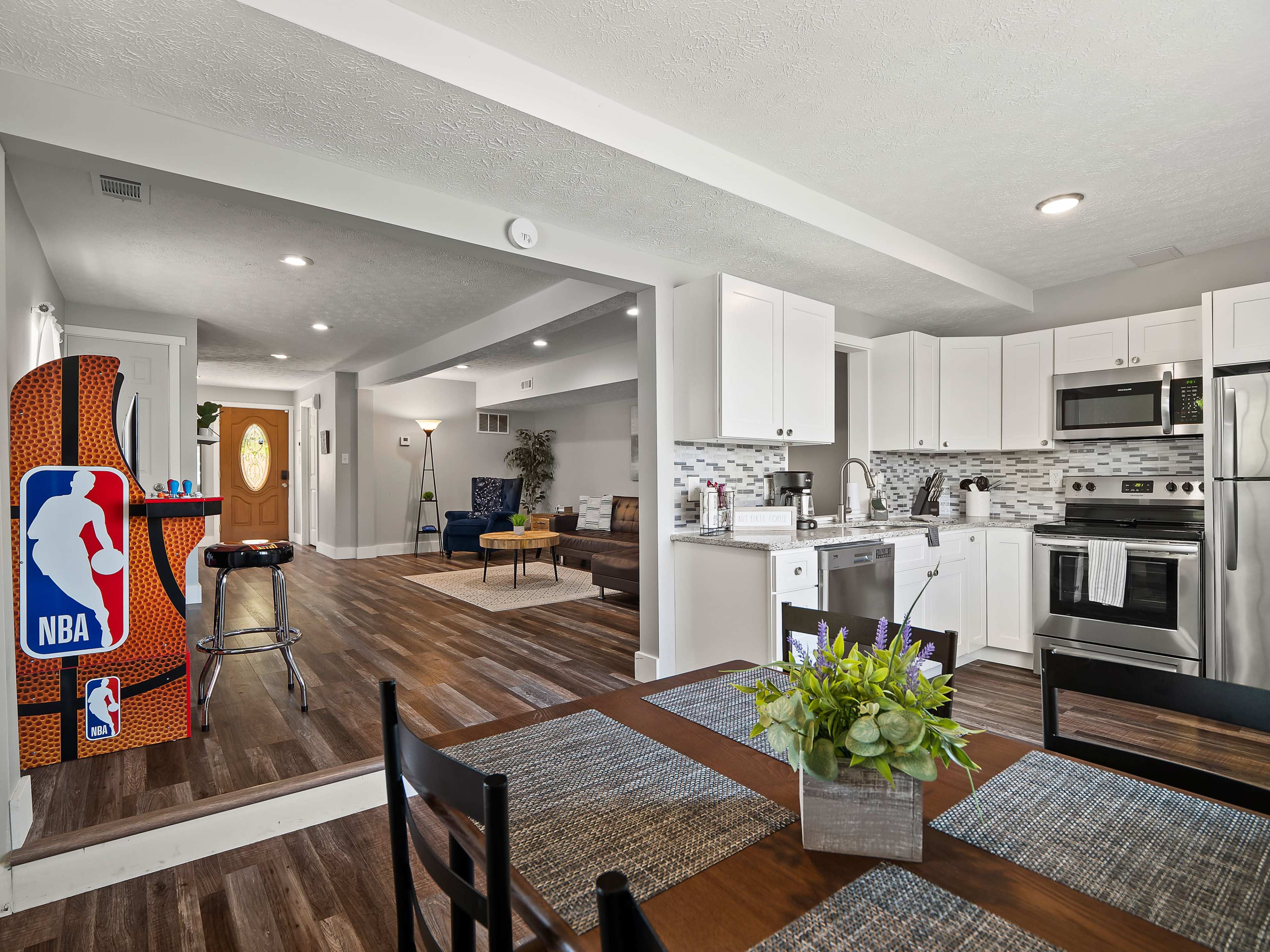 A modern kitchen and dining area with white cabinetry, stainless steel appliances, and a view into a living room featuring an NBA-themed arcade game.