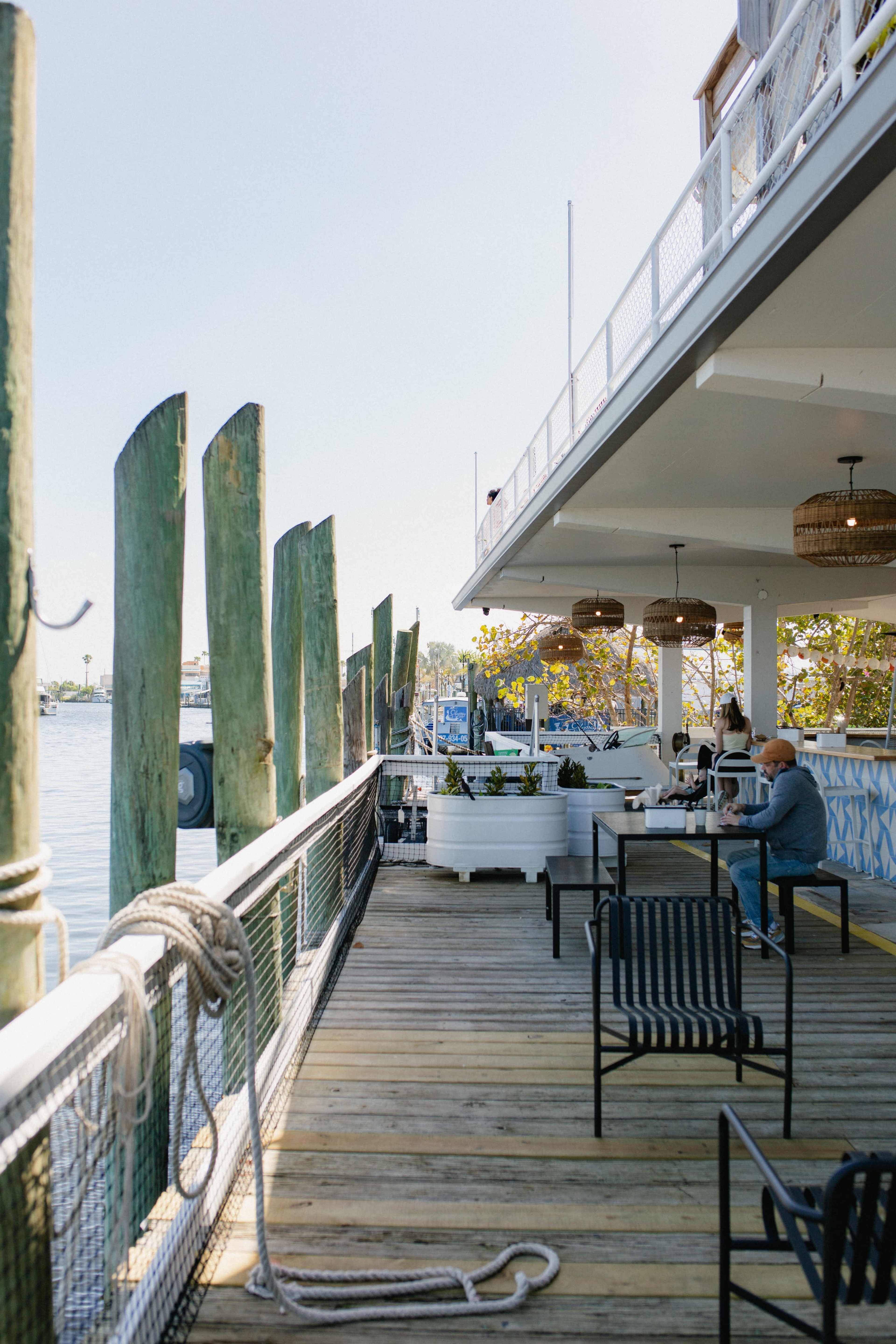 A wooden deck extends alongside a waterfront restaurant, with tables and chairs set up under a covered area and weathered green pilings along the edge.