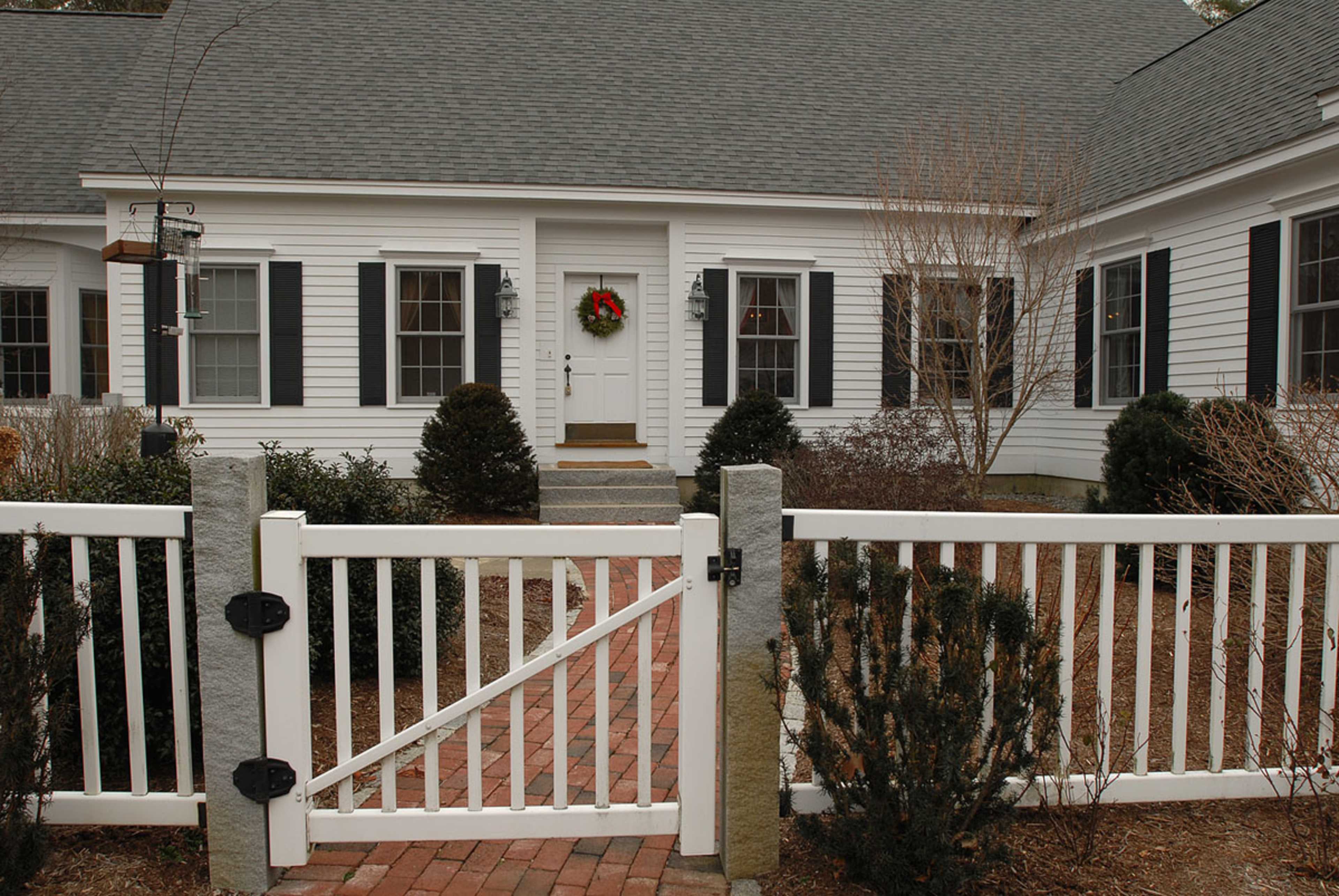 A white wooden fence with a gate leads to a house with a grey roof, featuring a wreath on the front door and landscaped gardens on either side.