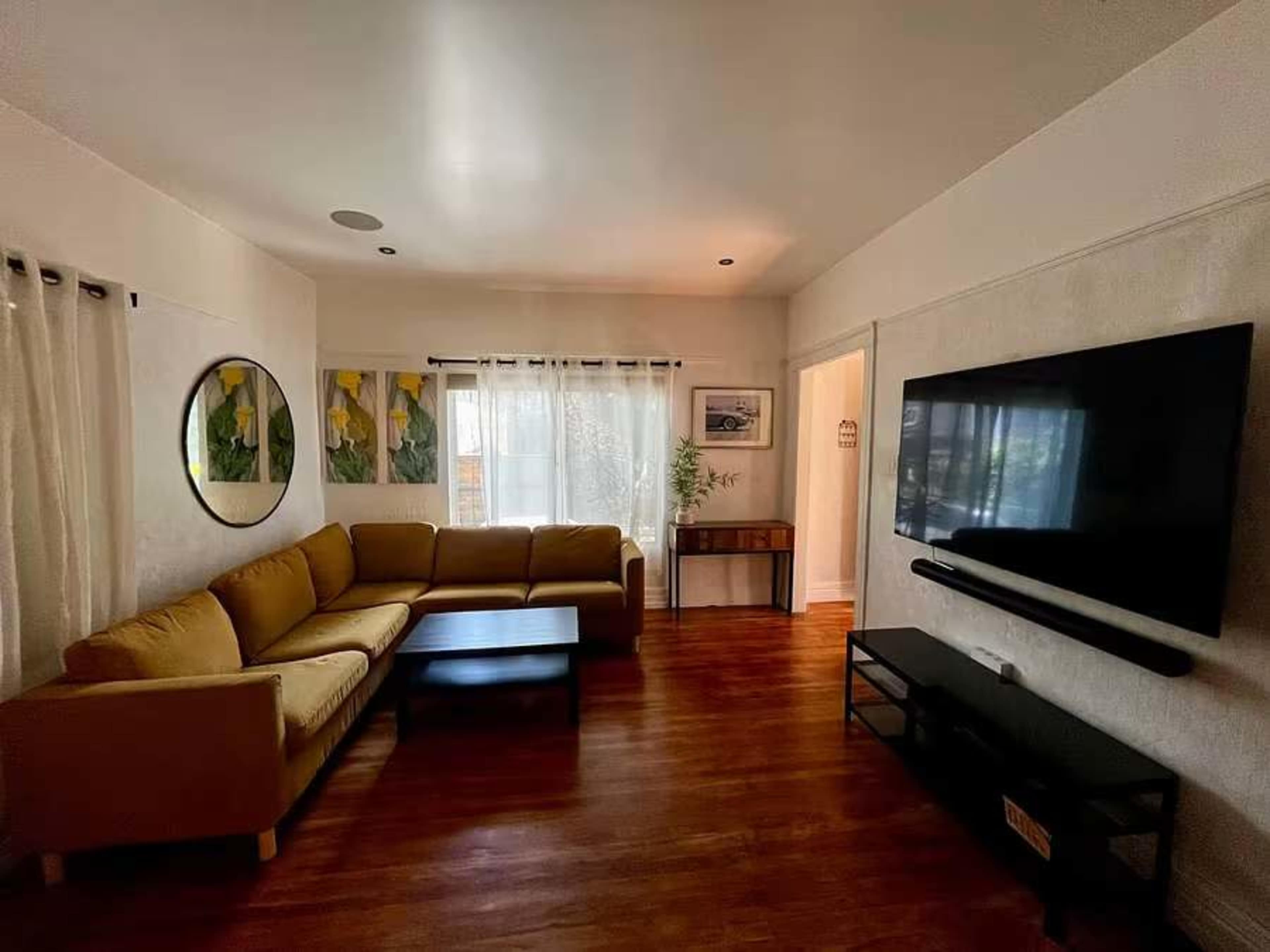 The living room features a large beige sectional sofa, a black coffee table, a wall-mounted television, and a window with sheer curtains, complemented by a circular wall mirror and framed artwork.