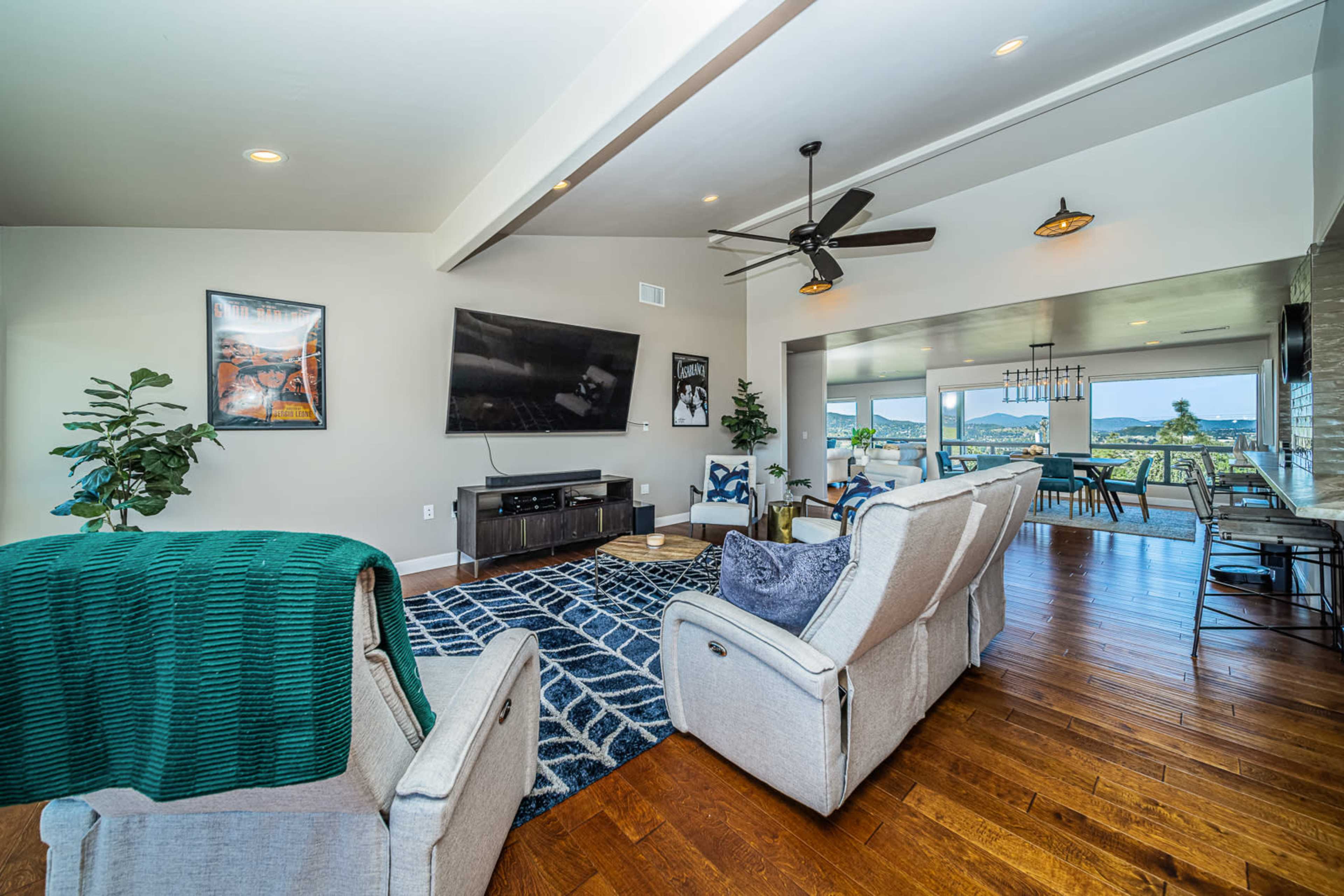 The image shows a modern living room featuring a television mounted on a wall, comfortable seating, a patterned area rug, and large windows offering a view of the outdoors.