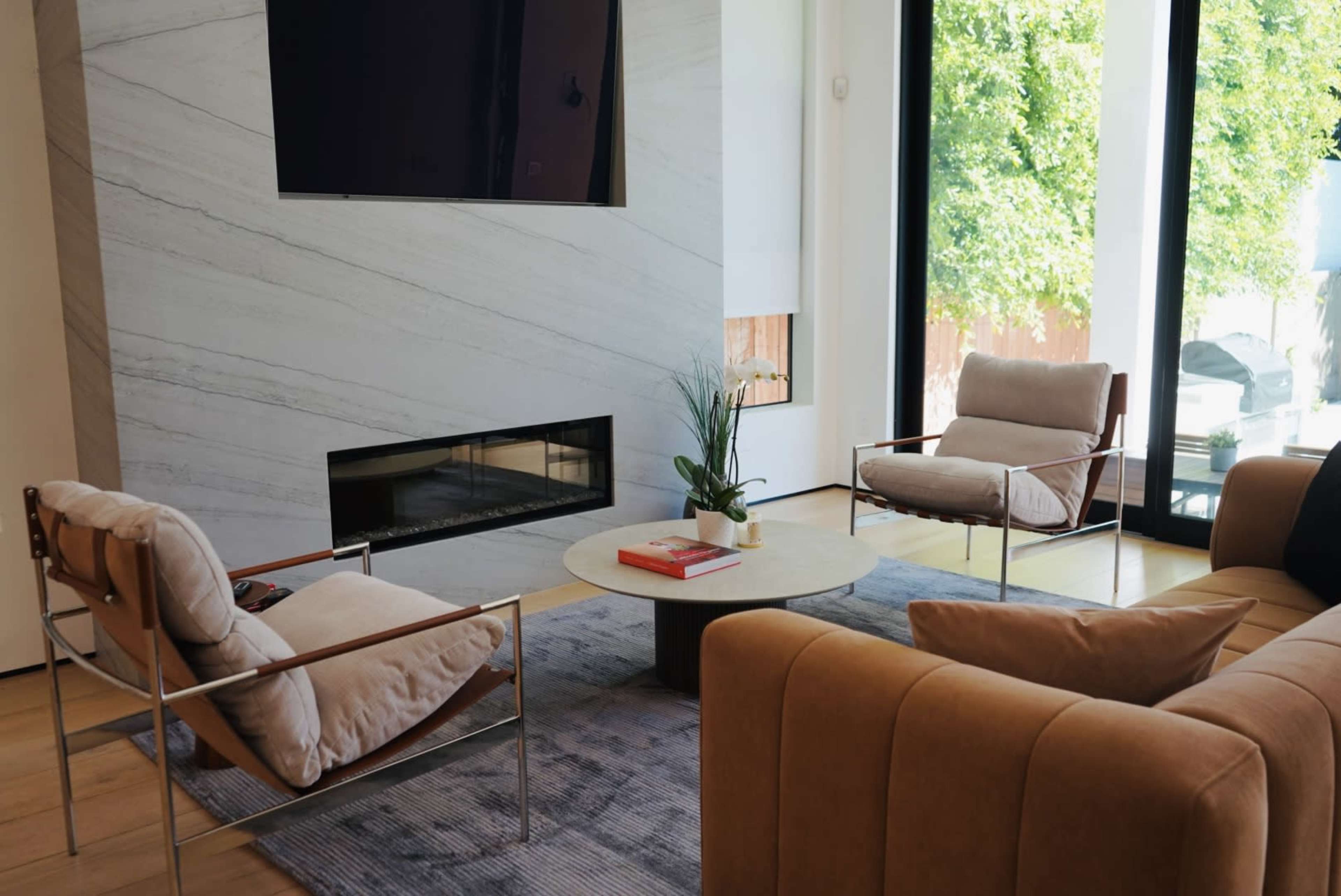 The image shows a modern living room with a marble fireplace, a circular coffee table, and two armchairs arranged around a small area rug.
