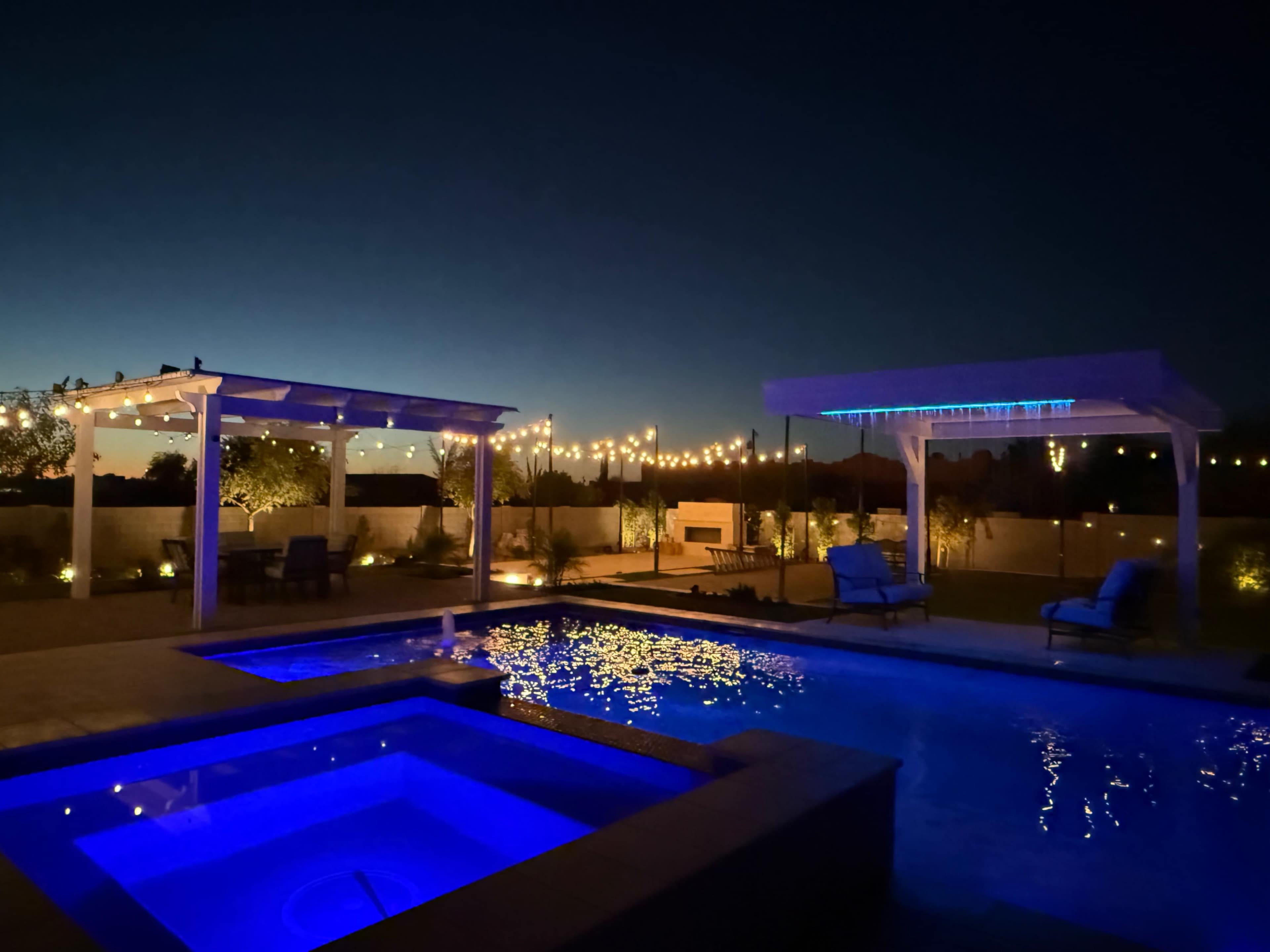 The image shows a well-lit outdoor pool area at night, featuring a swimming pool with blue lighting, a hot tub, and string lights illuminating the surrounding landscape.