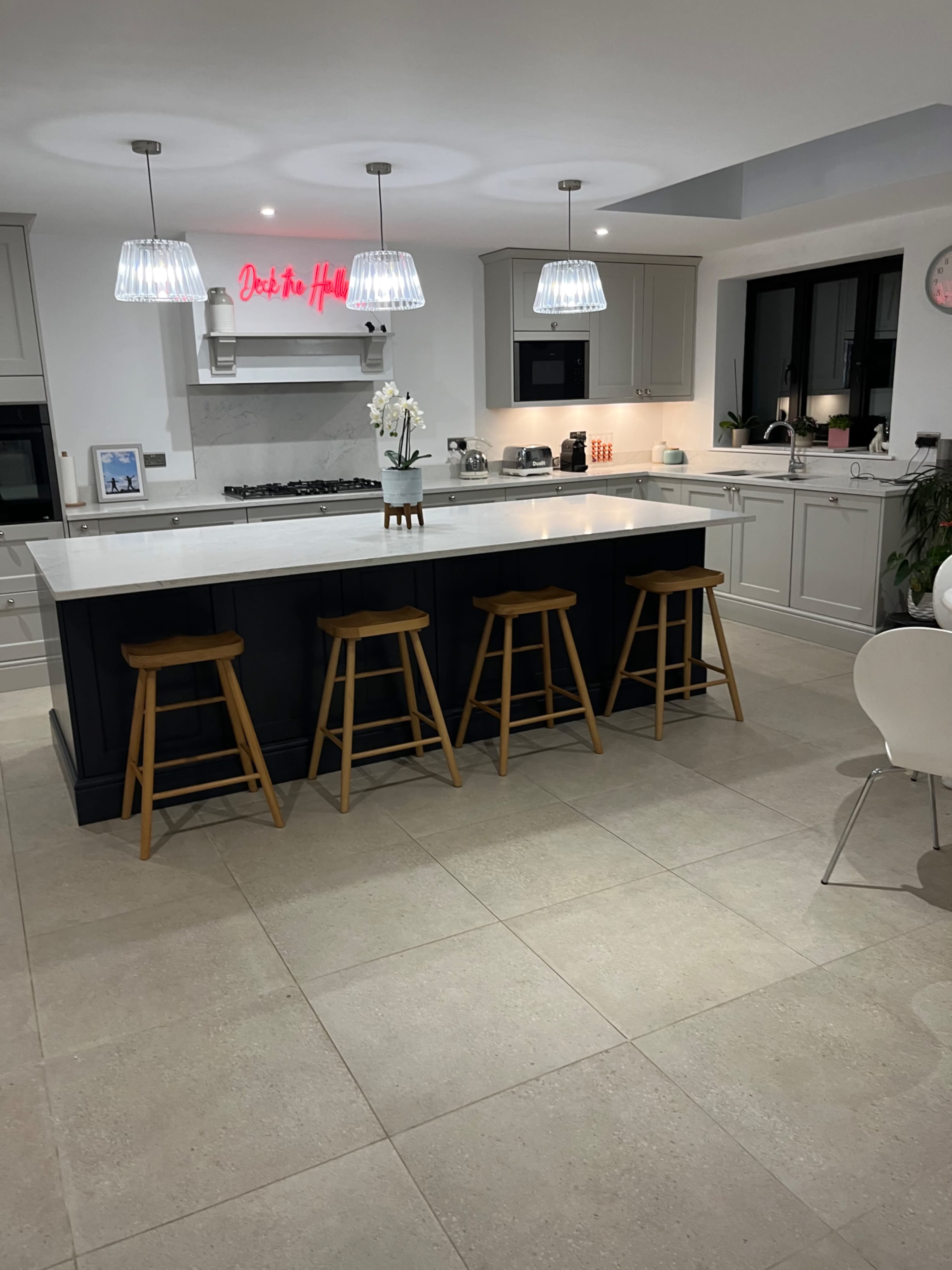 A modern kitchen features a large island with four wooden stools, pendant lights overhead, and a minimalist design with light cabinetry and a marble countertop.