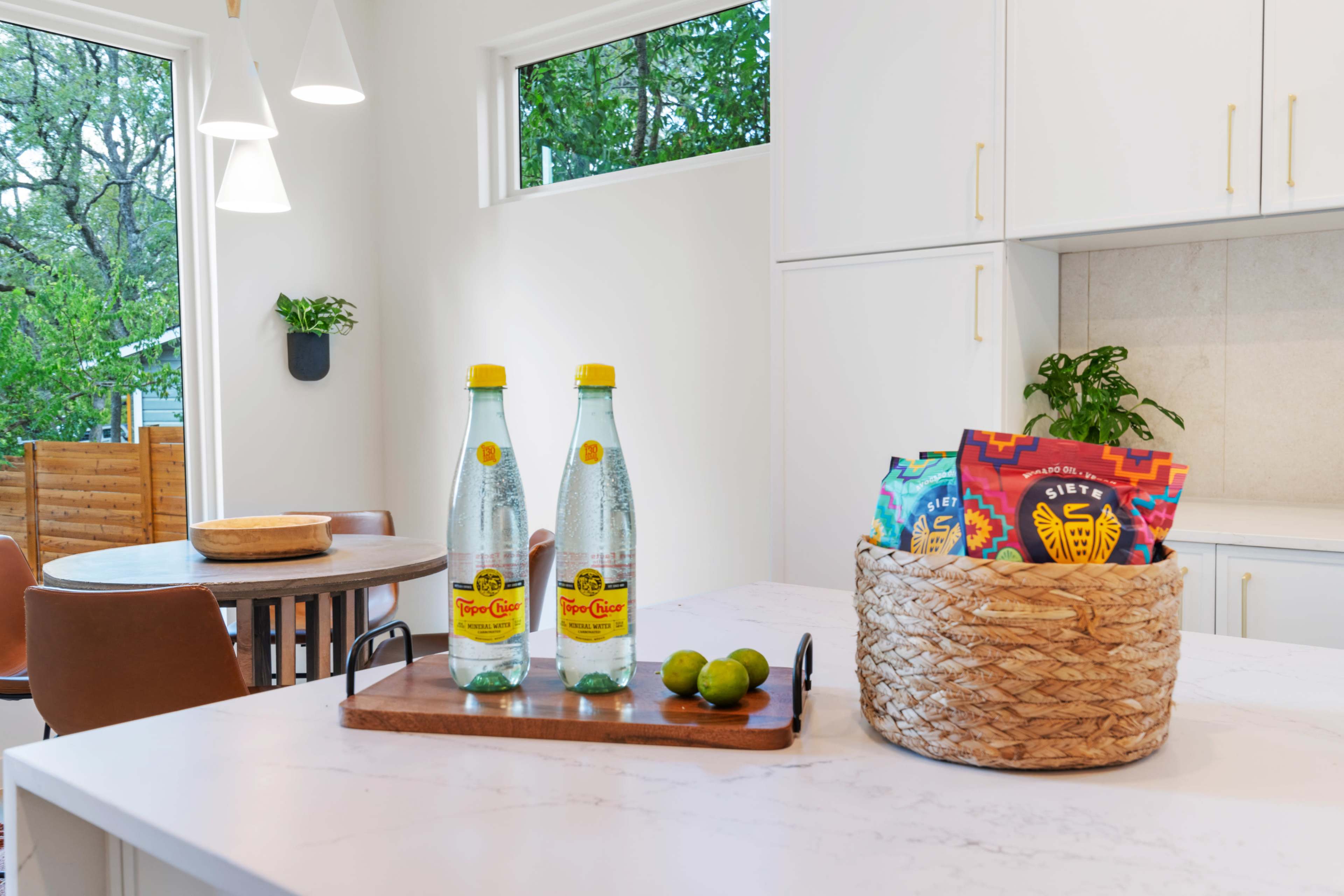 The image shows a modern kitchen with two bottles of sparkling water, a bowl of limes, and a woven basket containing colorful snack packages on a marble countertop.