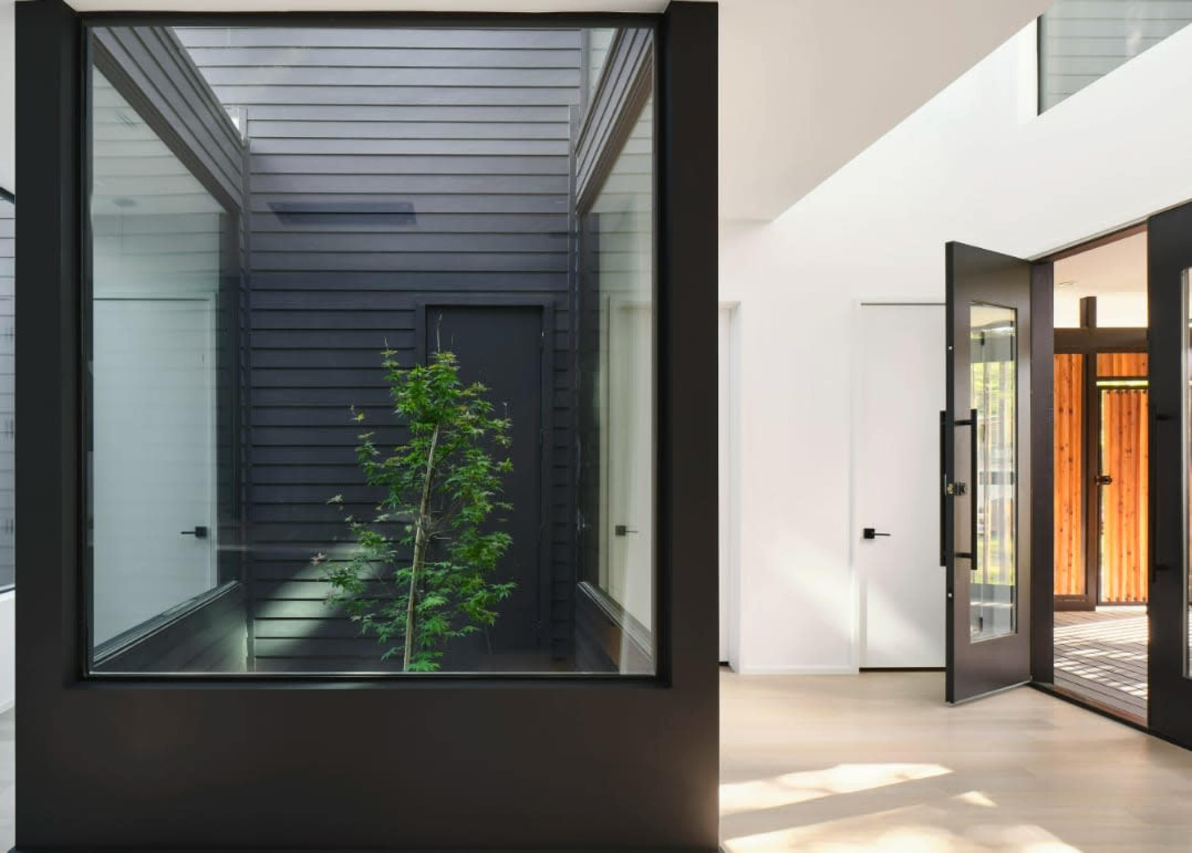 The image shows a modern interior space featuring large glass windows with a view of a small tree in a courtyard and doors leading to an outdoor area.